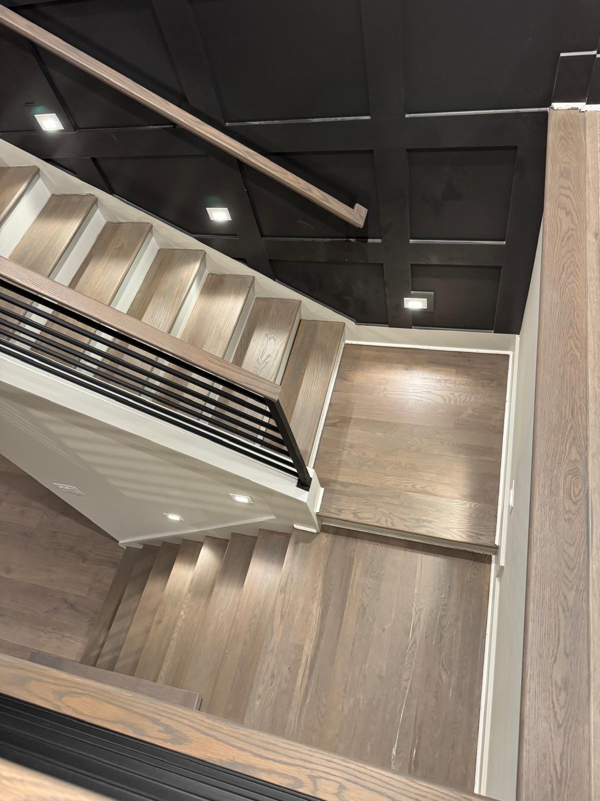 Interior view of a modern staircase with wooden steps, black railing, black paneled wall, and wooden flooring at the landing.