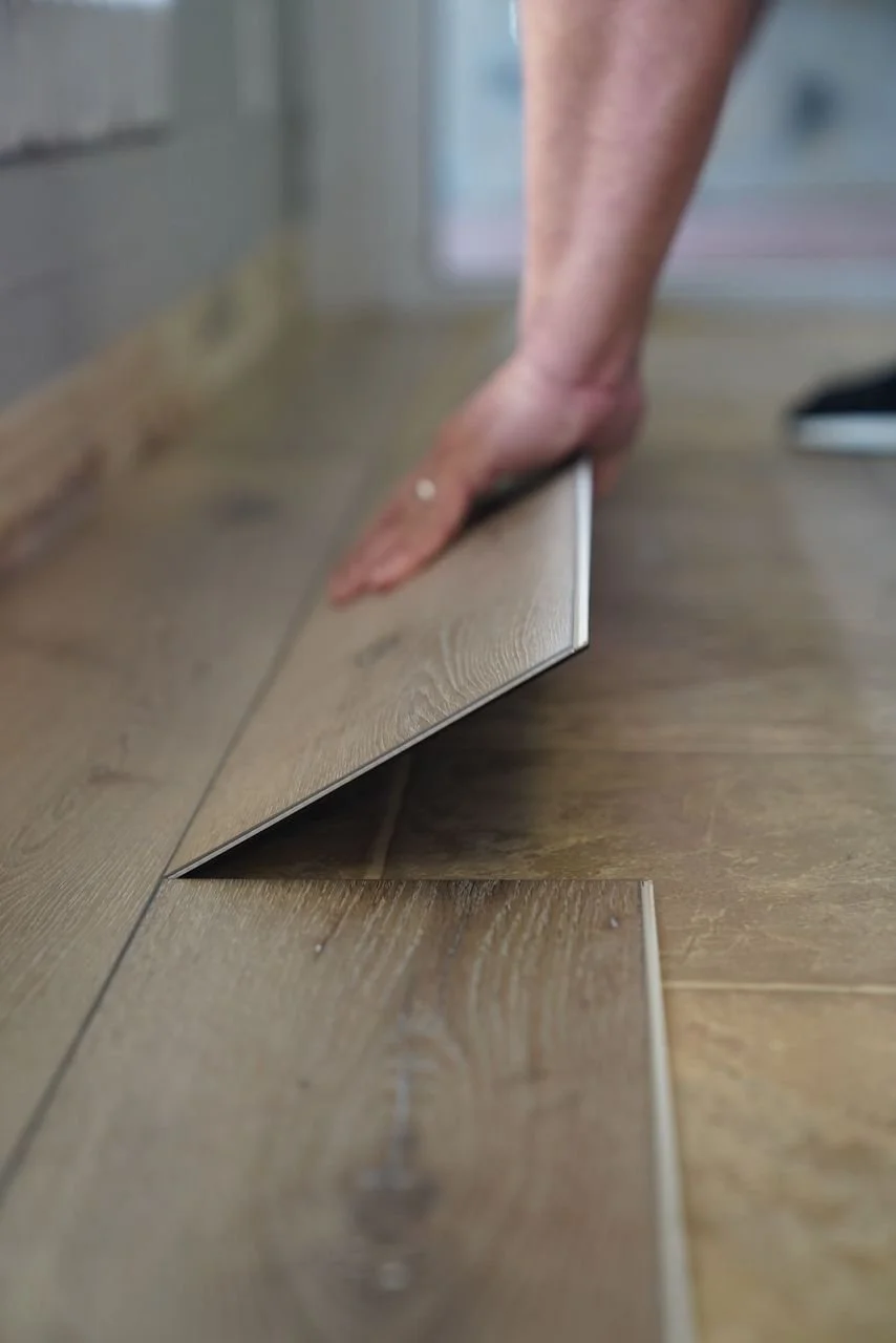 A person's hand installing a wooden floor tile, with the tile slightly lifted and being pressed into place on the floor.