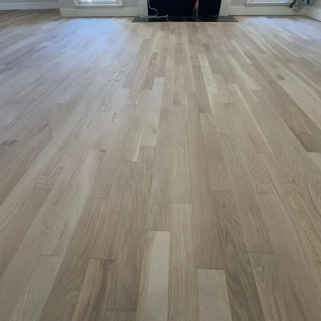 Light-colored hardwood floor in a room with large windows and a vent on the floor near the wall.