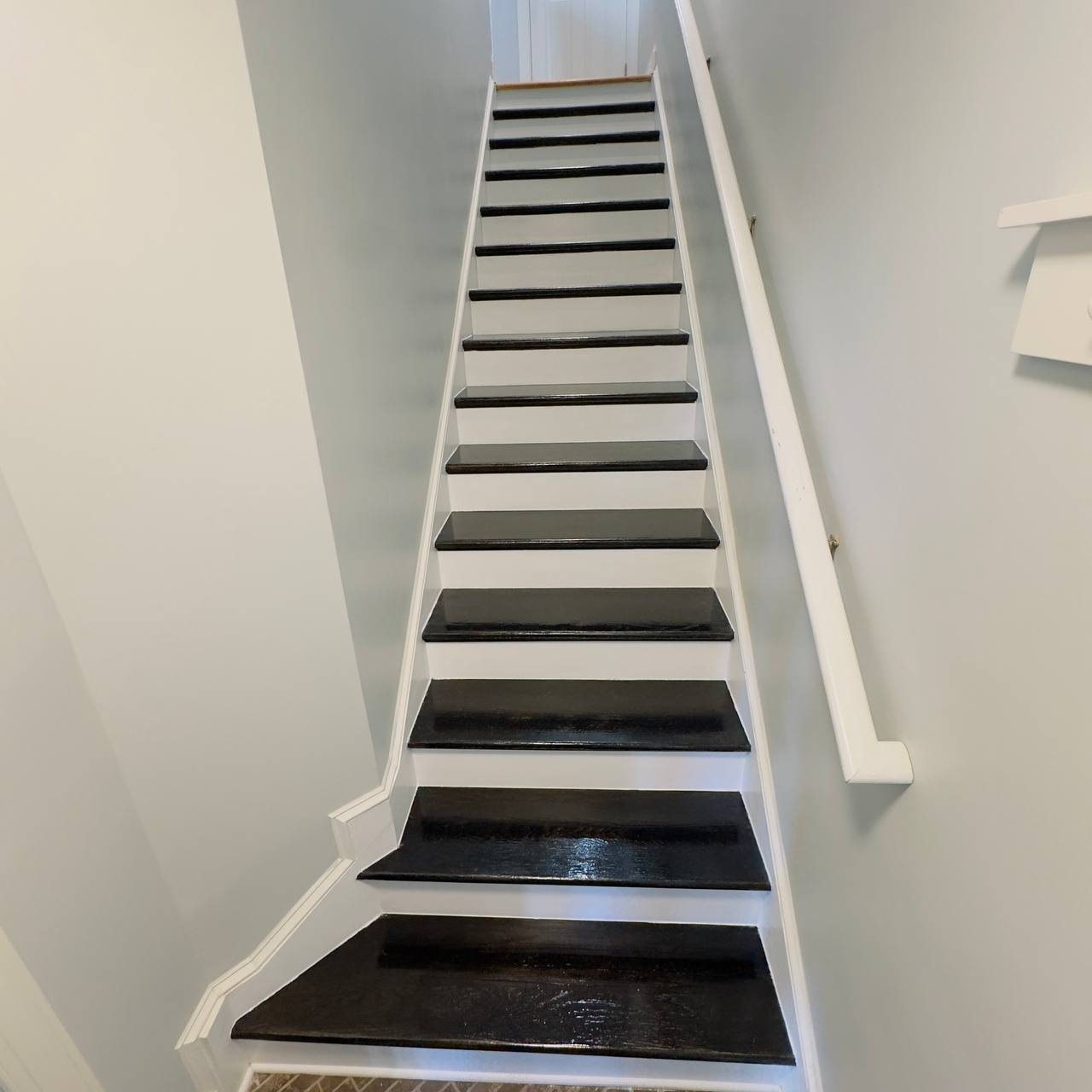 Image of a staircase with black painted treads, white risers, white walls, and a white handrail on the right side.