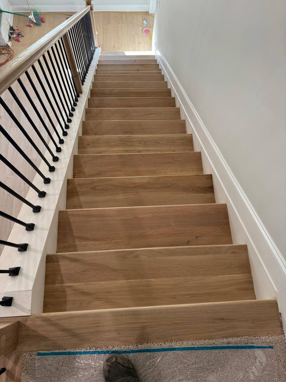Wooden stairs leading downward with black metal railing on the left and a wall on the right, part of a home's interior with construction or renovation materials visible at the top and side.
