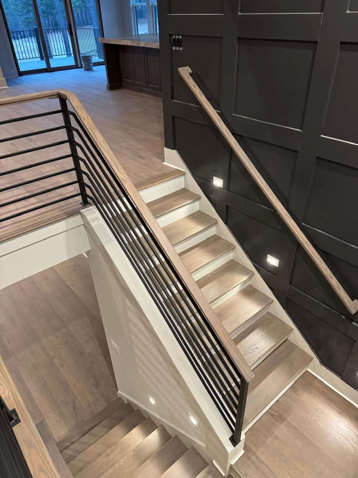 Interior view of a staircase in a modern home with wooden steps, a black metal and wood handrail, and black wall paneling with built-in lighting, leading from a lower to an upper level with wood flooring.