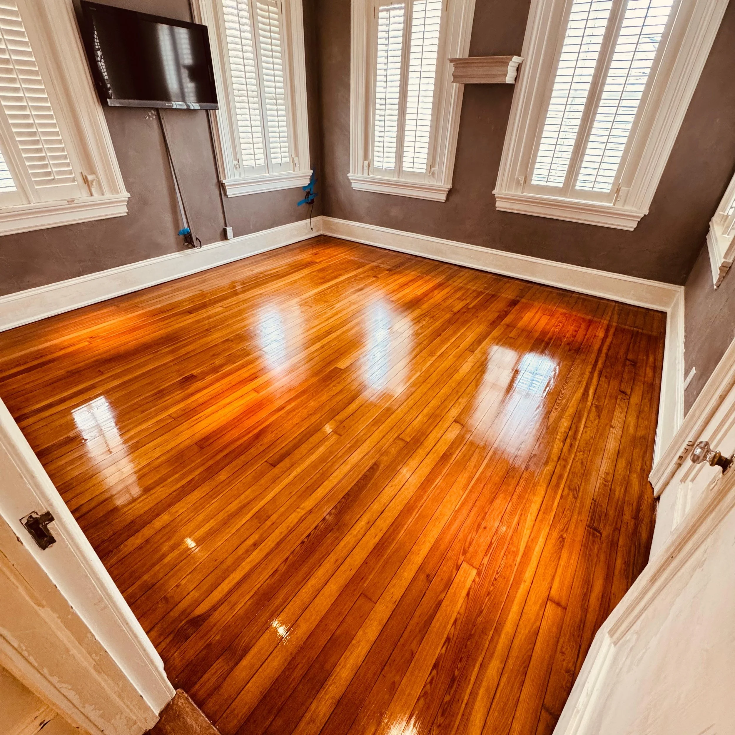 Empty room with hardwood flooring, brown walls, white trimmed windows with shutters, and a mounted television on the wall.