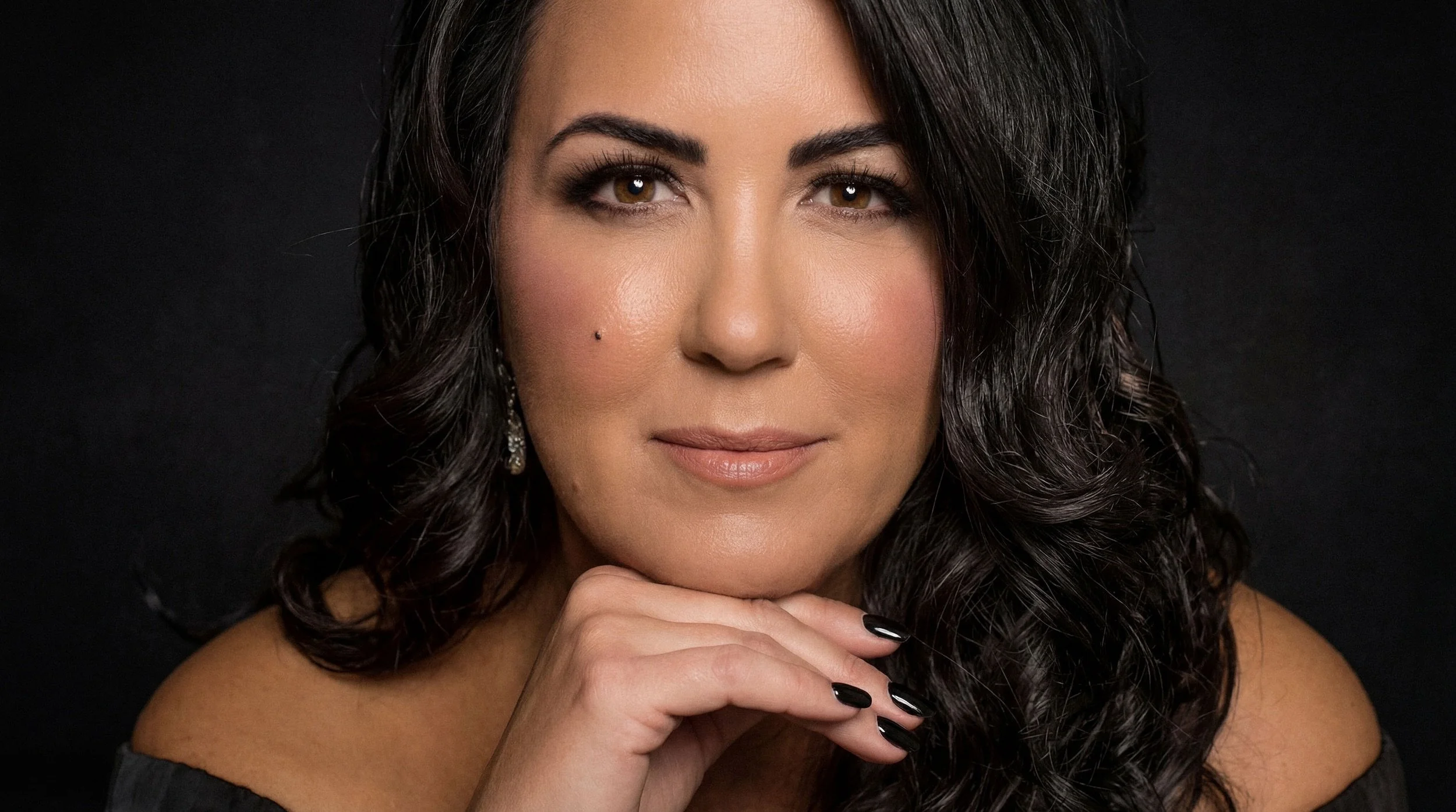 A woman with long dark curly hair, brown eyes, and fair skin looks directly at the camera with a subtle smile. She rests her chin on her hand, which has black painted nails, and wears a black off-the-shoulder top and dangling earrings against a dark background.