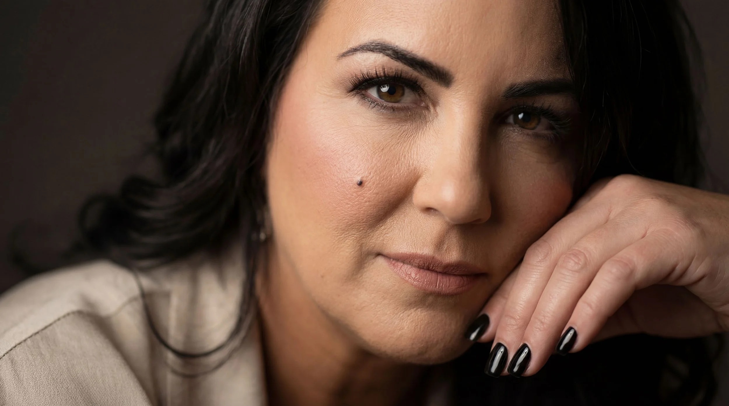 Close-up portrait of a woman with dark hair and brown eyes, resting her chin on her hand, with makeup and a small beauty mark near her cheek.