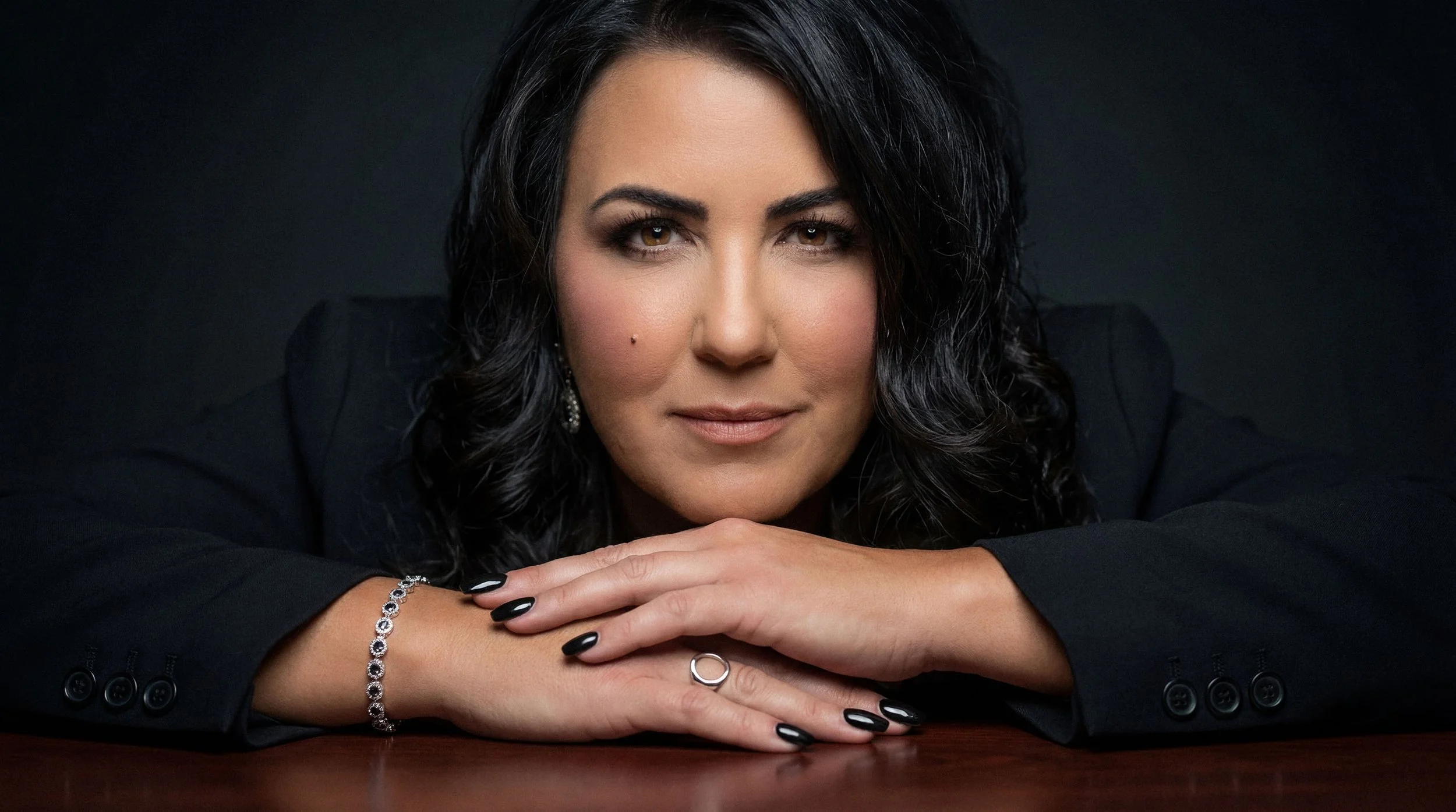 Close-up portrait of a woman with black hair resting her chin on her crossed arms, wearing a black blazer and jewelry, against a dark background.