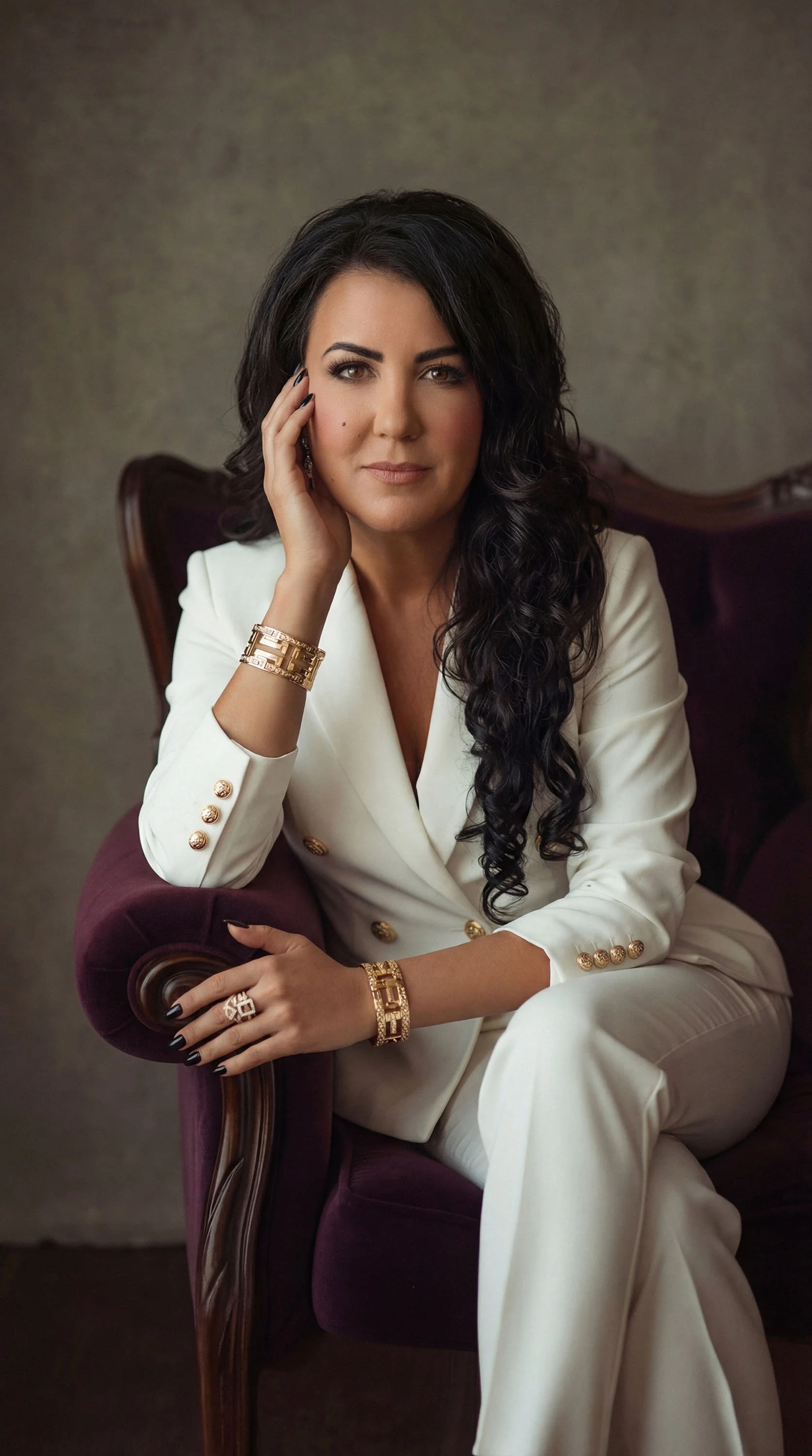 A woman with long dark curly hair seated on a dark purple vintage armchair, wearing a white suit with gold buttons, accessorized with gold jewelry, and resting her chin on her hand.