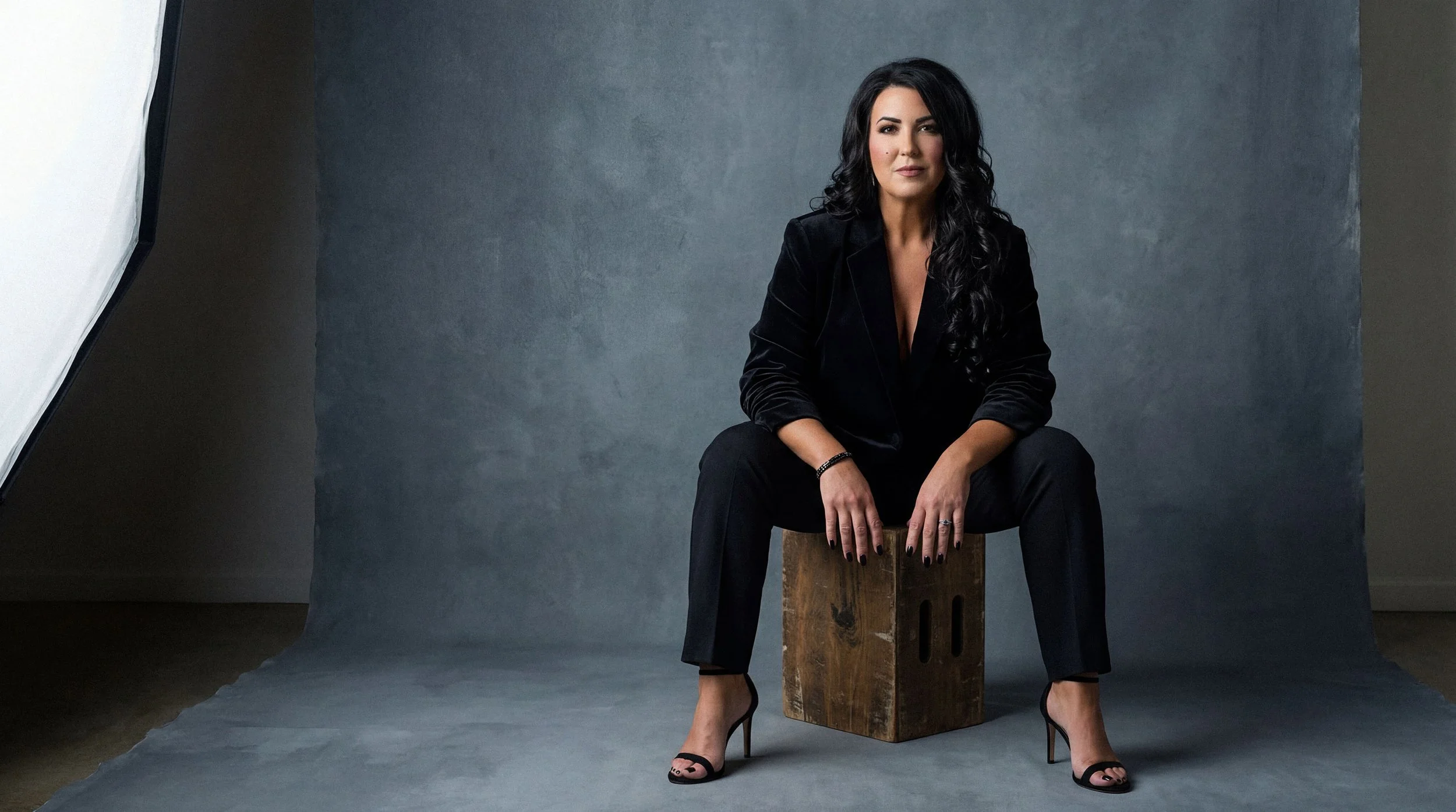 A woman with long curly black hair, wearing a black velvet blazer and black pants, seated on a wooden box against a gray backdrop in a photography studio.