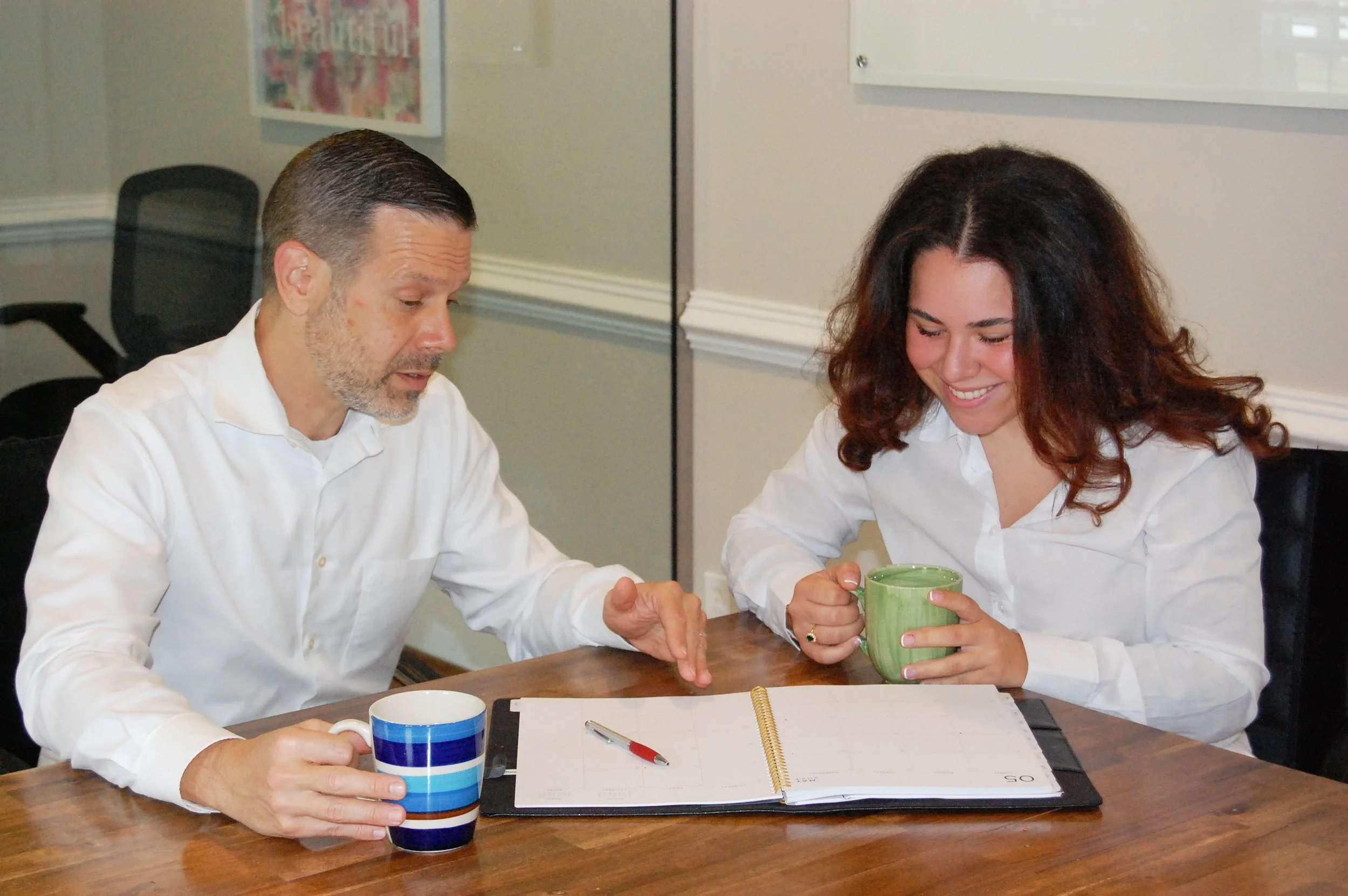 A man and woman sitting at a wooden table, looking at a planner with a red pen on it. The man holds a blue striped mug, and the woman holds a green mug, both smiling.