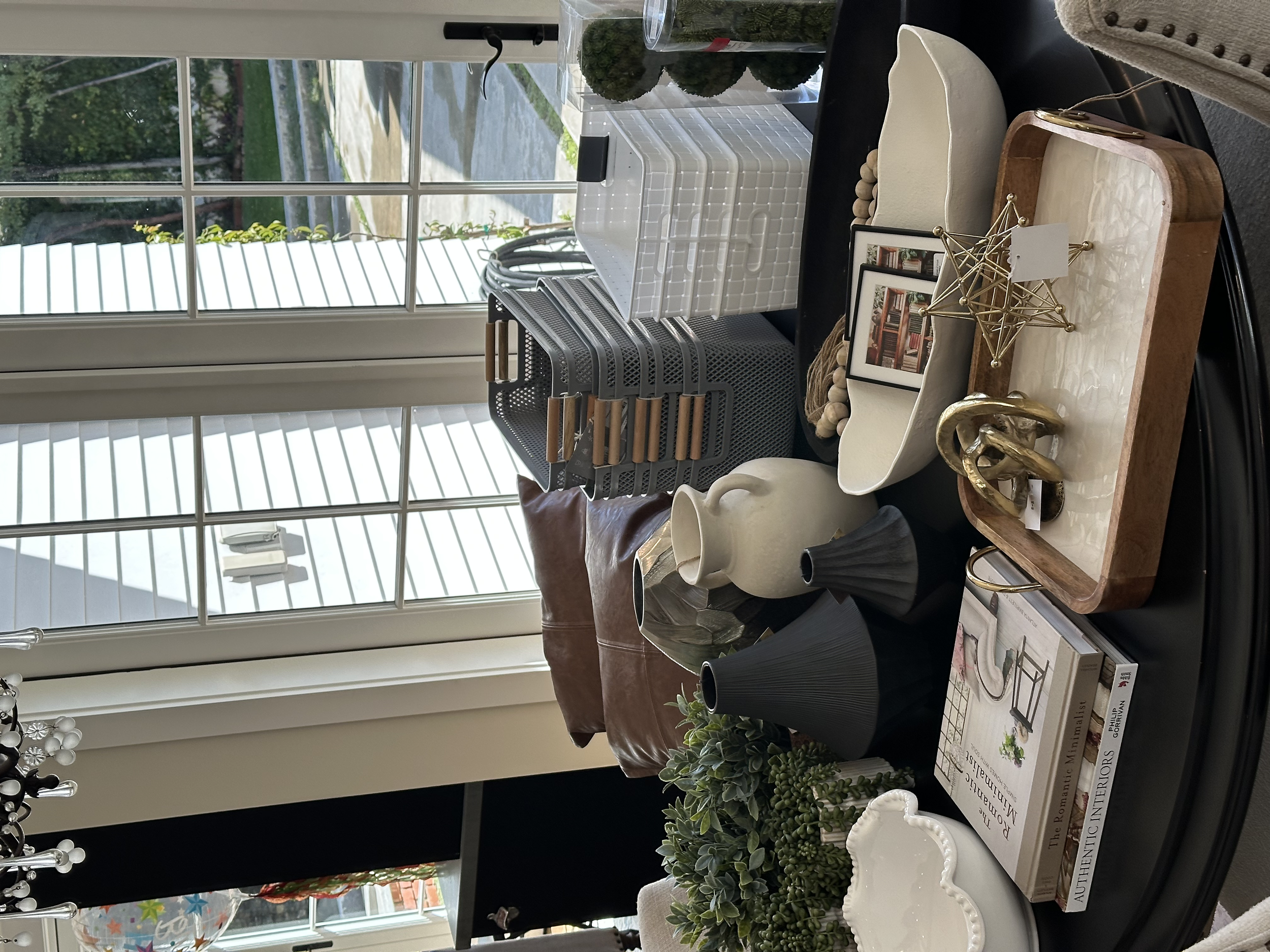 Decorative tabletop display with vases, picture frames, plants, books, and sculptures near a window with white blinds.
