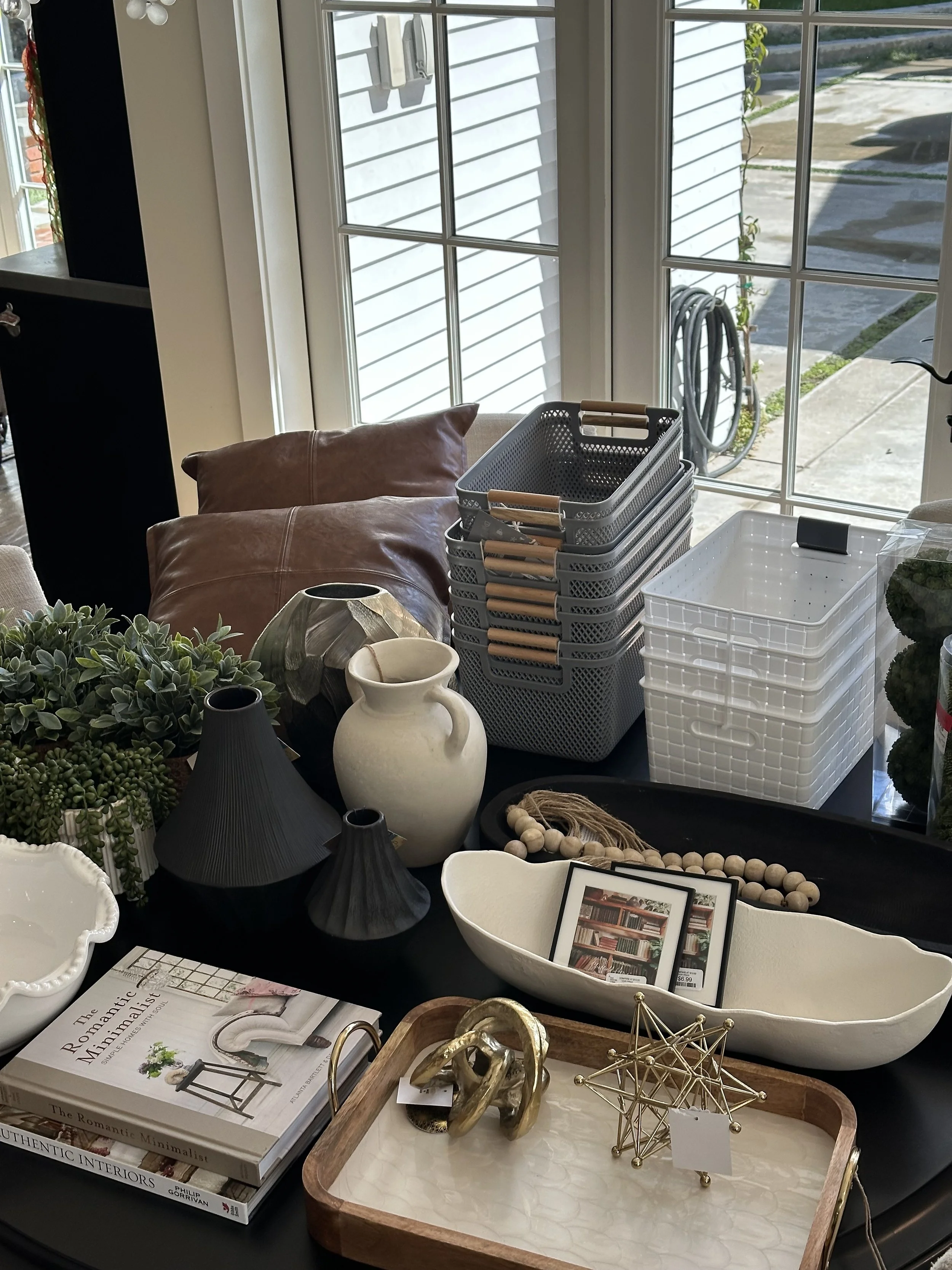 Decorative objects on a table near a window, including stackable baskets, vases, lamps, framed pictures, books, and various ornamental items.