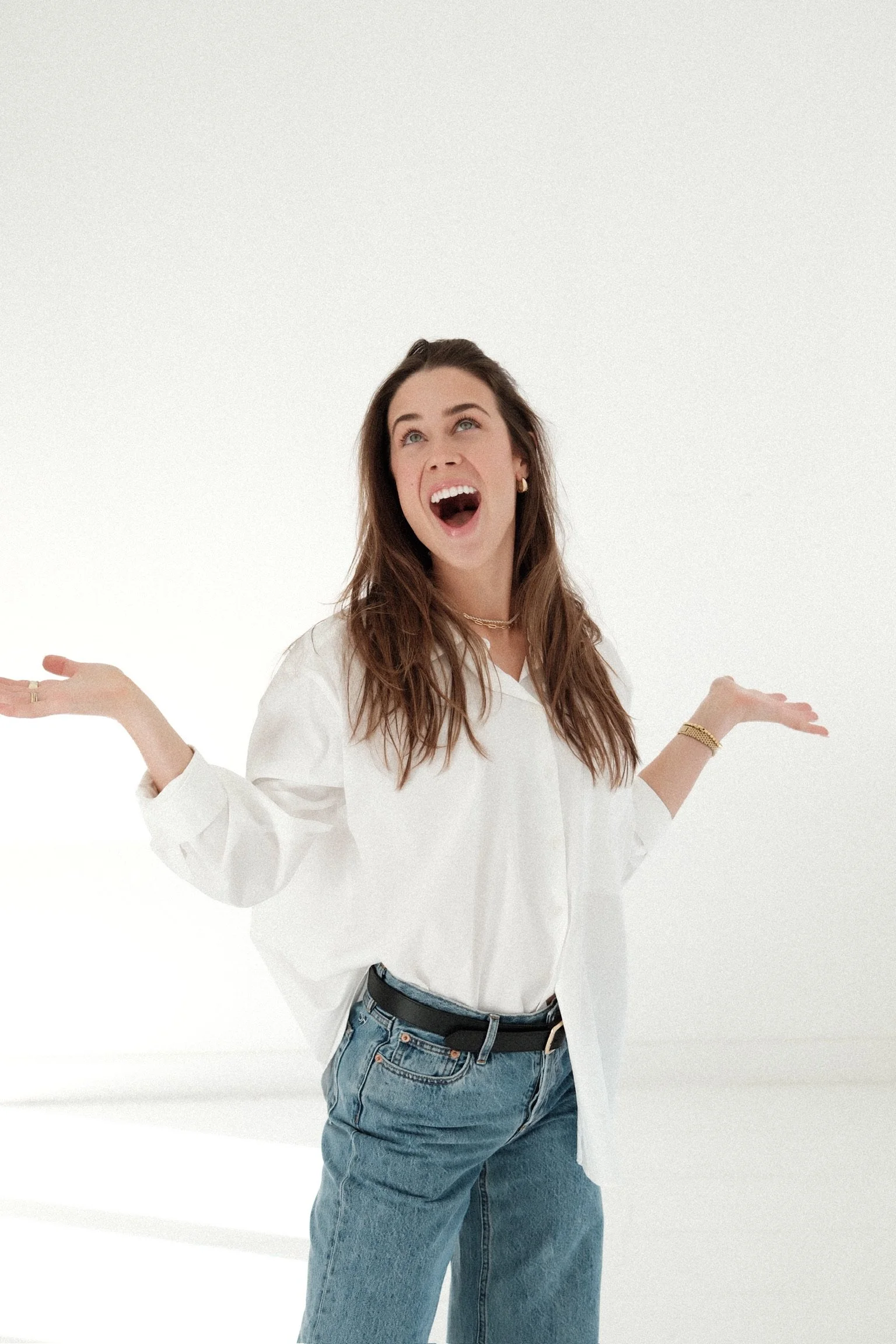 A young woman with long brown hair, wearing a white blouse and blue jeans, standing with her arms outstretched and smiling in a white studio setting.