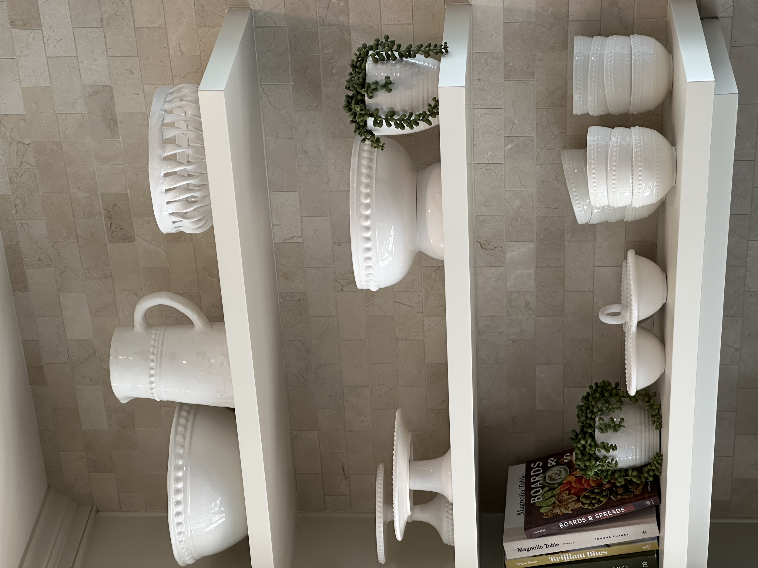 White ceramic dishware set including a mug, cake stands, vases, and bowls on a white shelf. There are small potted plants and cookbooks on the shelf as well, with a beige tiled wall in the background.