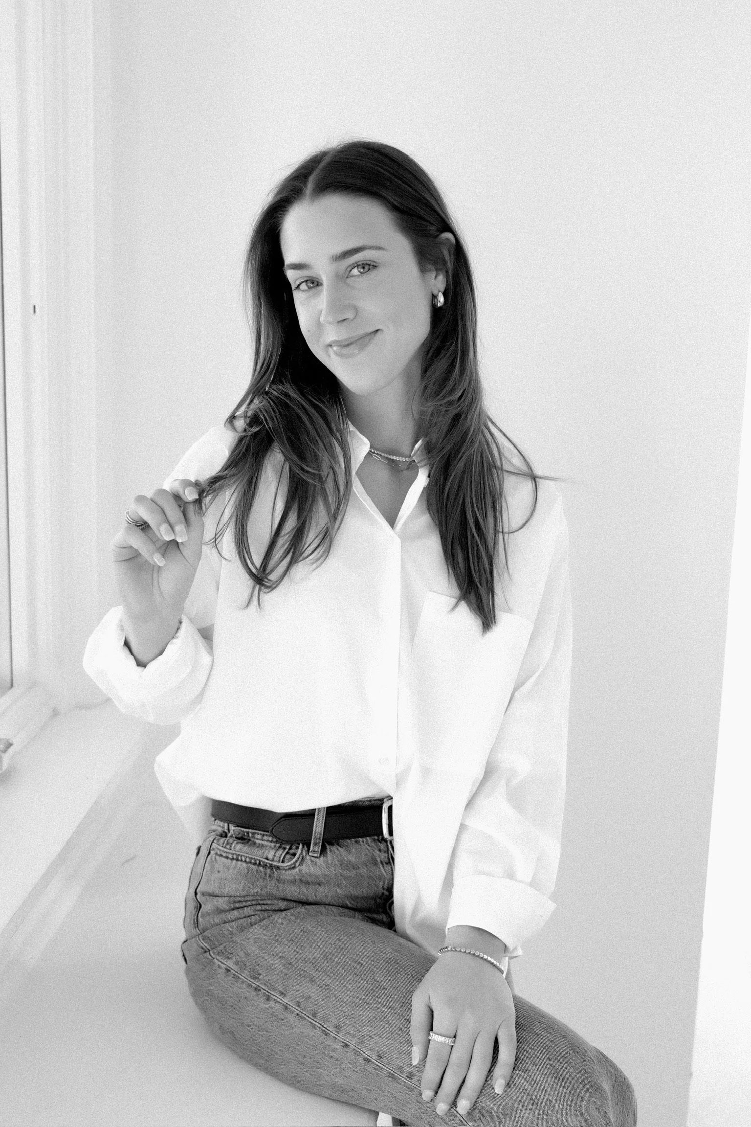A young woman with long dark hair sitting on a windowsill, wearing a white blouse, jeans, and jewelry, smiling at the camera, in black and white.