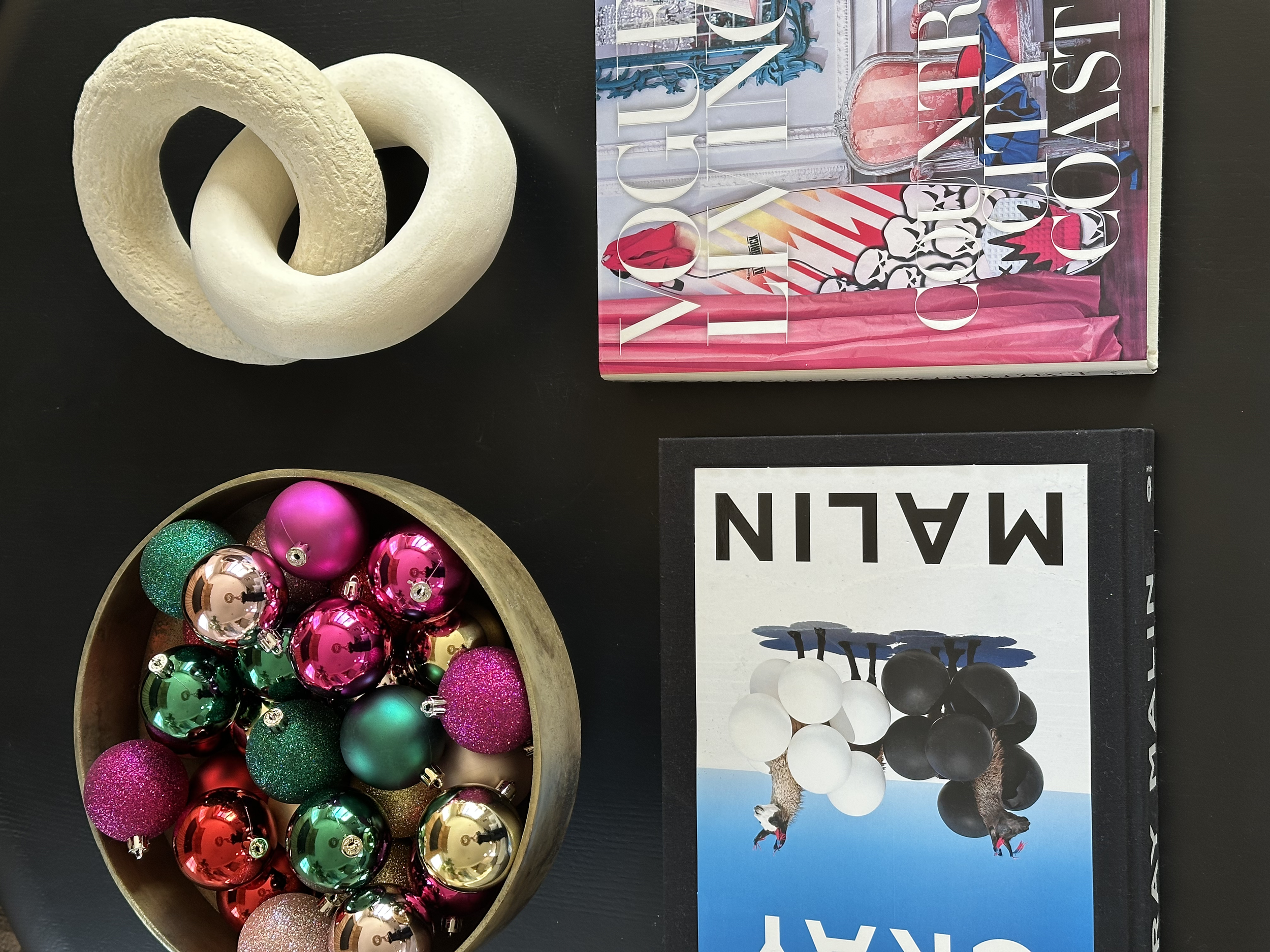 A black table holding two books, a wooden bowl filled with colorful Christmas ornaments, and two decorative torus-shaped objects. The books are labeled 'VOGUE LONDON' and 'MALIN'. The ornaments in the bowl are shiny in pink, green, gold, and red, som
