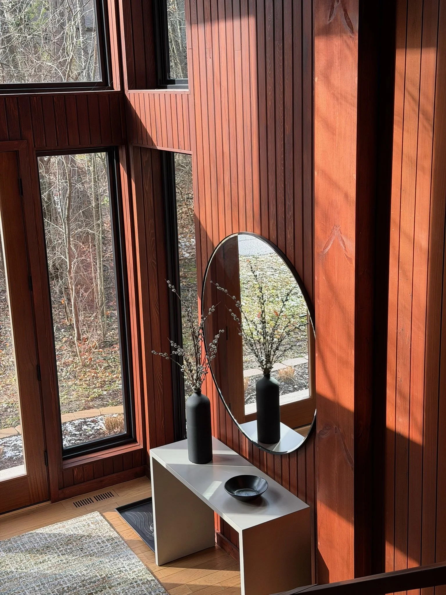 Interior view of a room with wooden panel walls, large windows showing an outdoor scene with trees, a white table with black vases and a mirror, and a patterned rug on the floor.