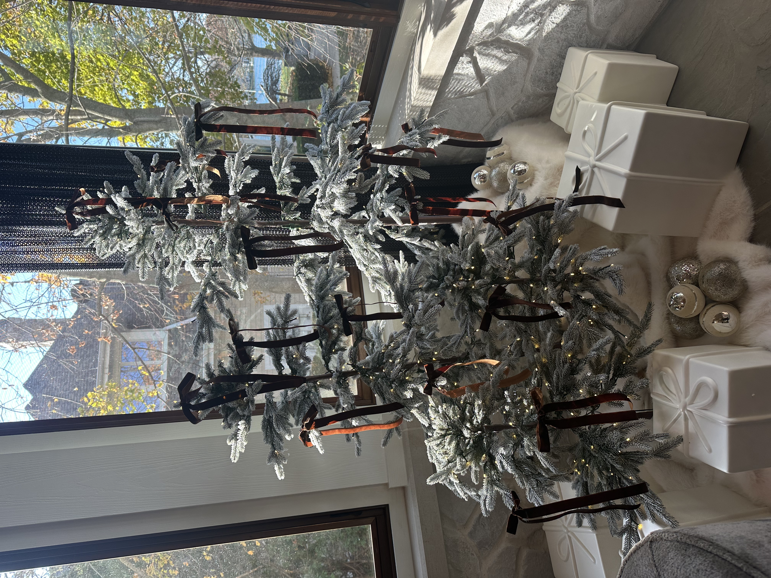 Decorated Christmas tree with silver and white ornaments, ribbons, and fairy lights, positioned near large windows with a view of trees and houses outside.