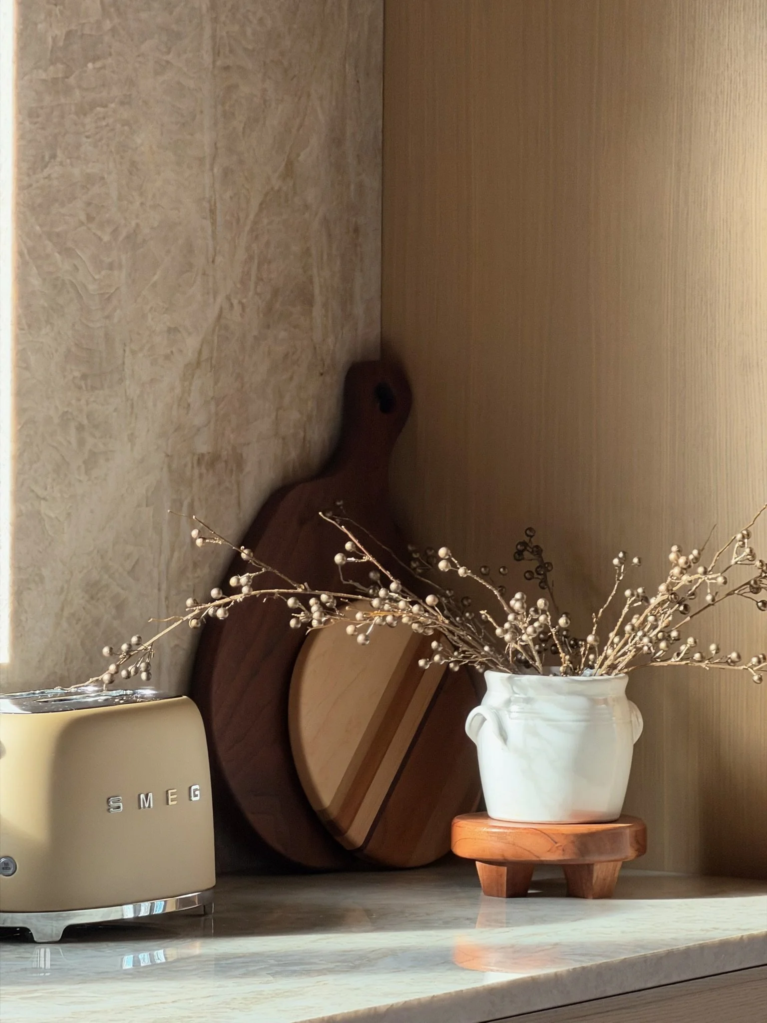 Kitchen counter with a SMEG toaster, wooden cutting boards, and a white vase with decorative branches, against a wall with wood paneling and tile.