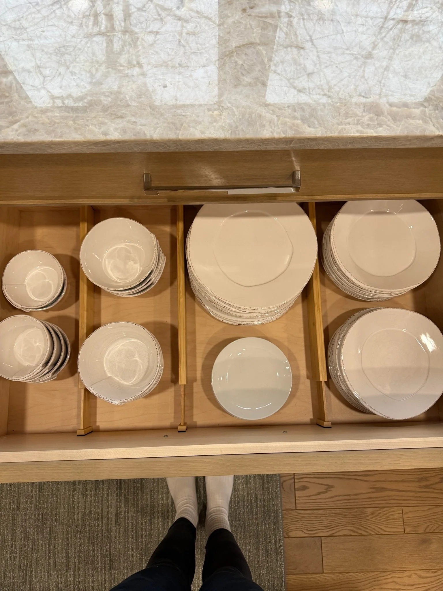 Open wooden drawer containing nested white plates of various sizes, with the photographer's legs and feet visible at the bottom, standing on carpeted and wooden floor.
