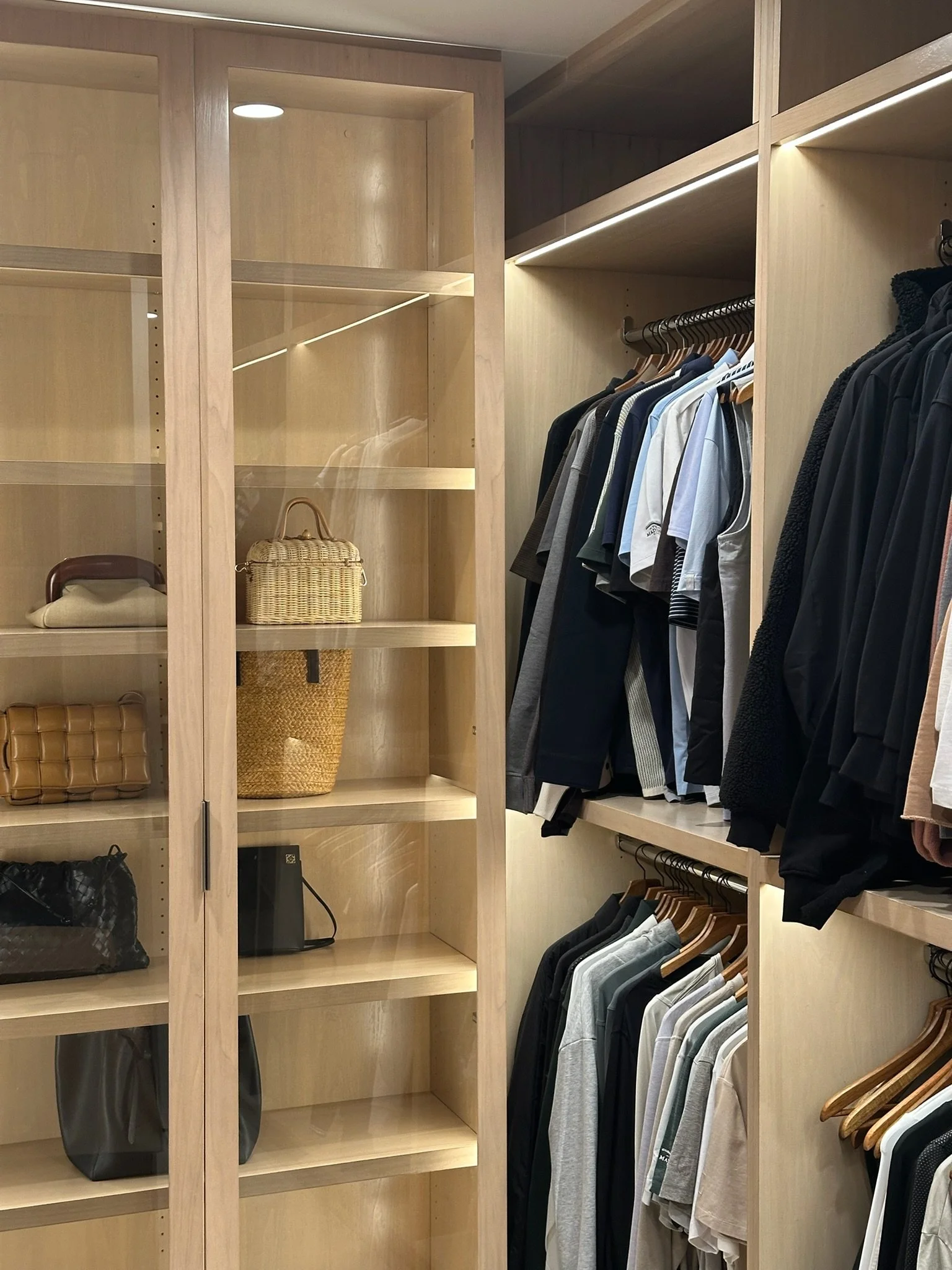 Closet with wooden shelves and hanging clothes, some handbags and a small electronic device on the shelves.