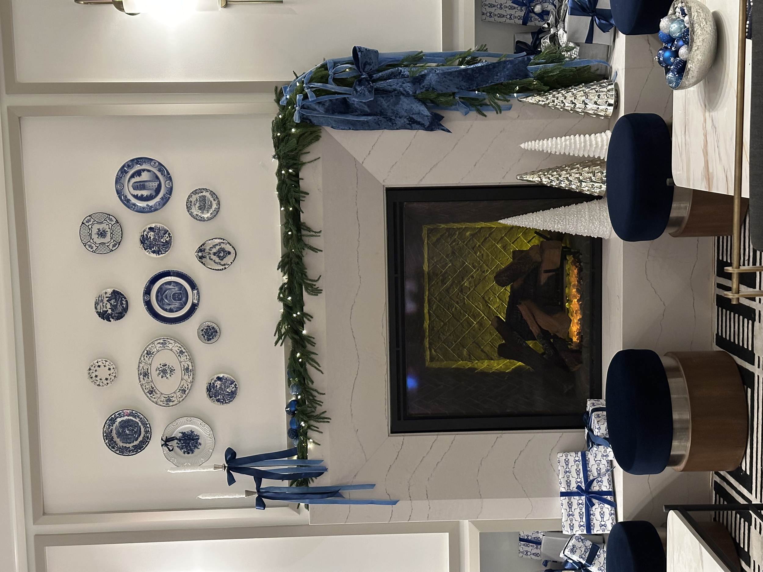 Living room with a fireplace decorated for Christmas with a green garland and blue ribbons. Blue and white Christmas ornaments and plates on the wall. Two wrapped presents on the table, and blue stools around. Christmas tree branches draped across th