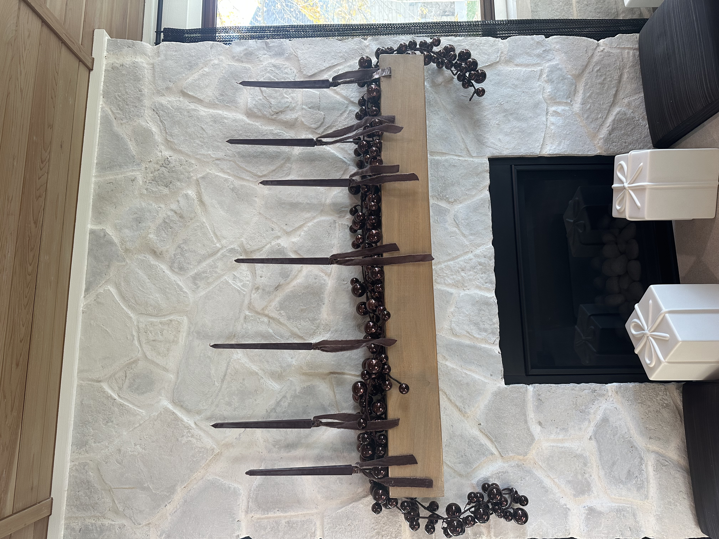 Decorative fireplace with a stone wall, adorned with black candles and a string of dark berries. Two white gift boxes with ribbons are placed on the right side.