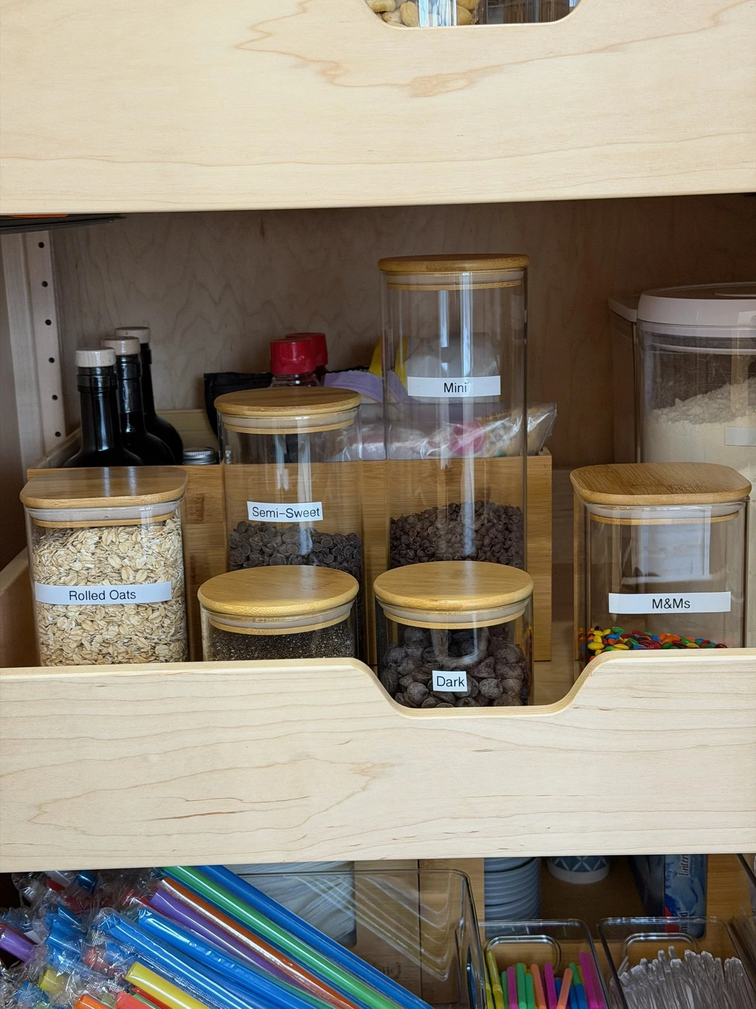 Organized pantry shelf with labeled containers of oats, semi-sweet chocolate chips, mini chocolate chips, dark chocolate, and M&Ms, with other kitchen items in the background.