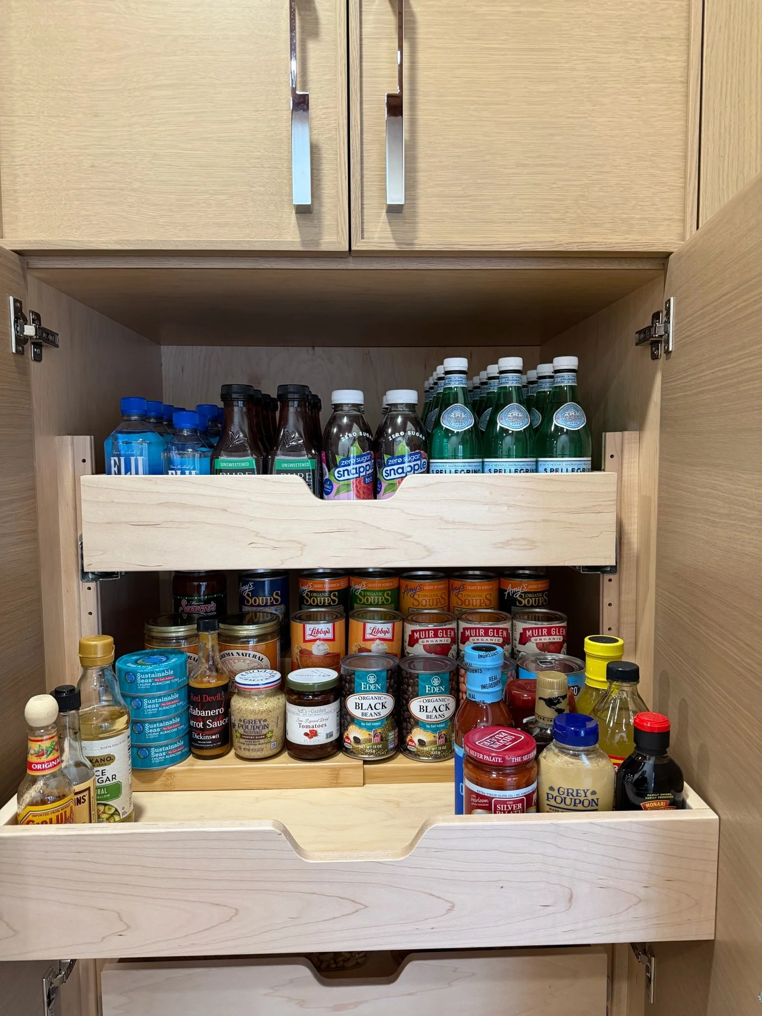 Open kitchen cabinet containing bottled water, flavored drinks, canned soups, beans, and condiments.