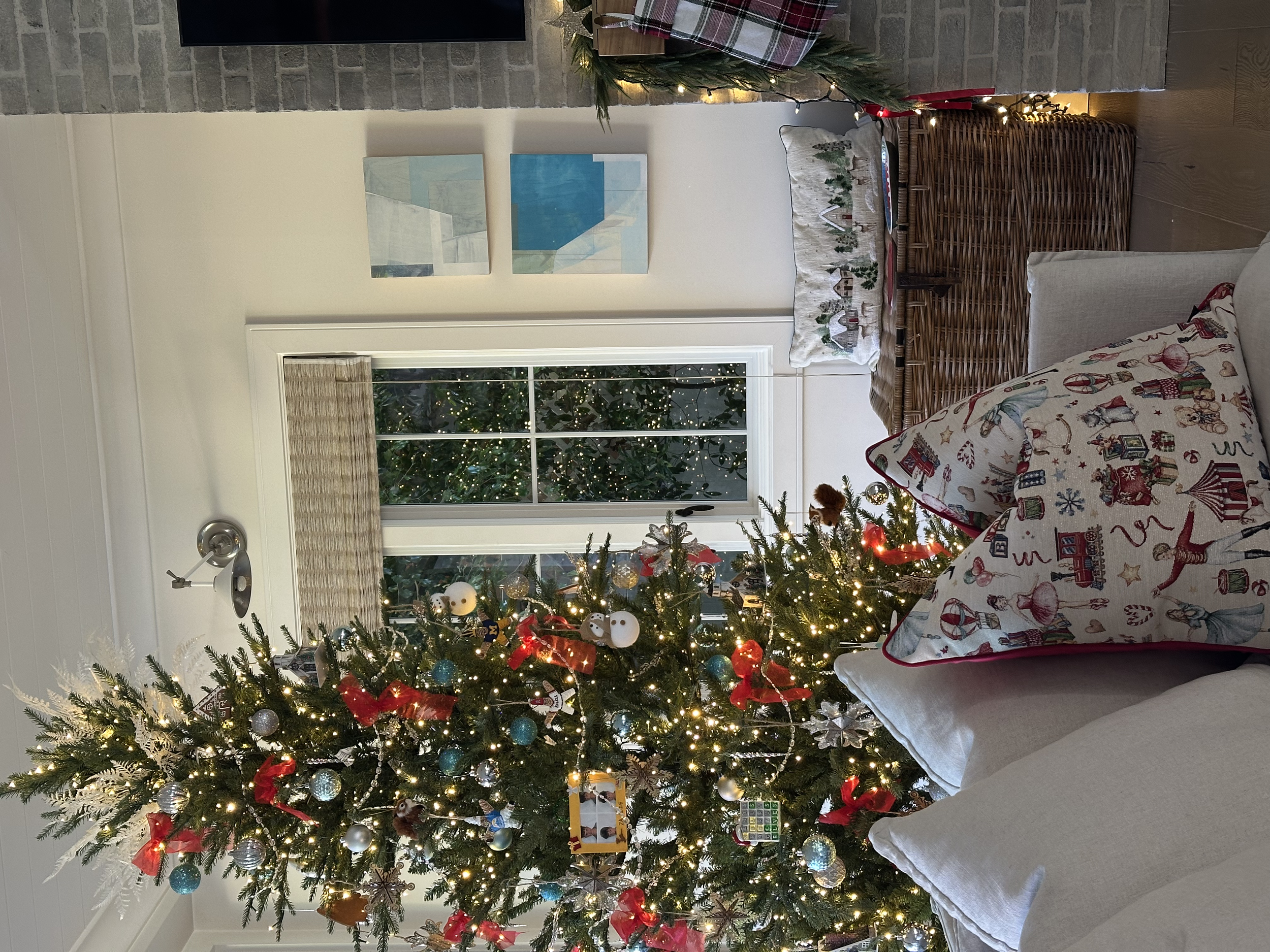A decorated Christmas tree with ornaments, lights, and ribbons in a cozy living room. A window showing outdoor greenery, a sofa with Christmas pillows, a wicker chest, and a wall with artwork are also visible.