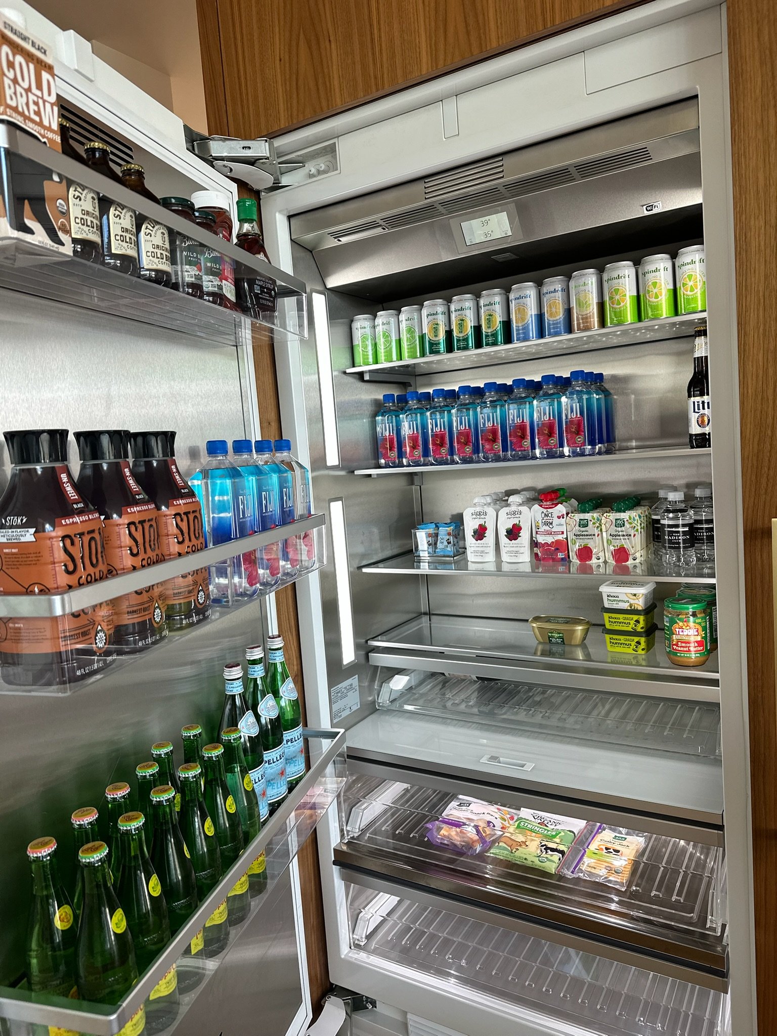 An open refrigerator with various beverages and food items inside, including bottled drinks on the shelves and some packaged food in the drawers.