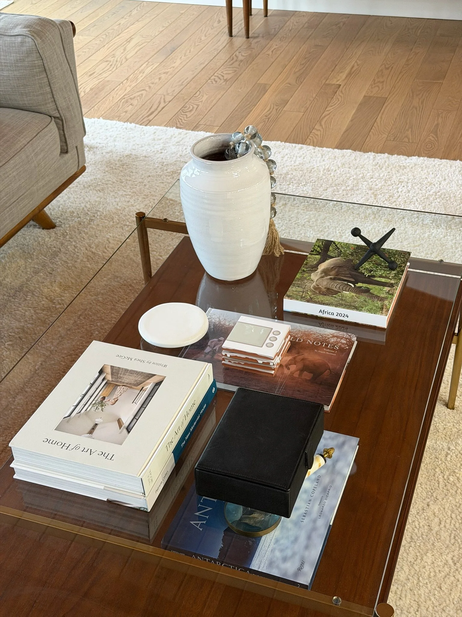 A glass-top coffee table with a white ceramic vase, a book titled "Africa 2024," a small white dish, a set of small books, a black jewelry box, and various other books and decorative items on a wooden and beige rug background.