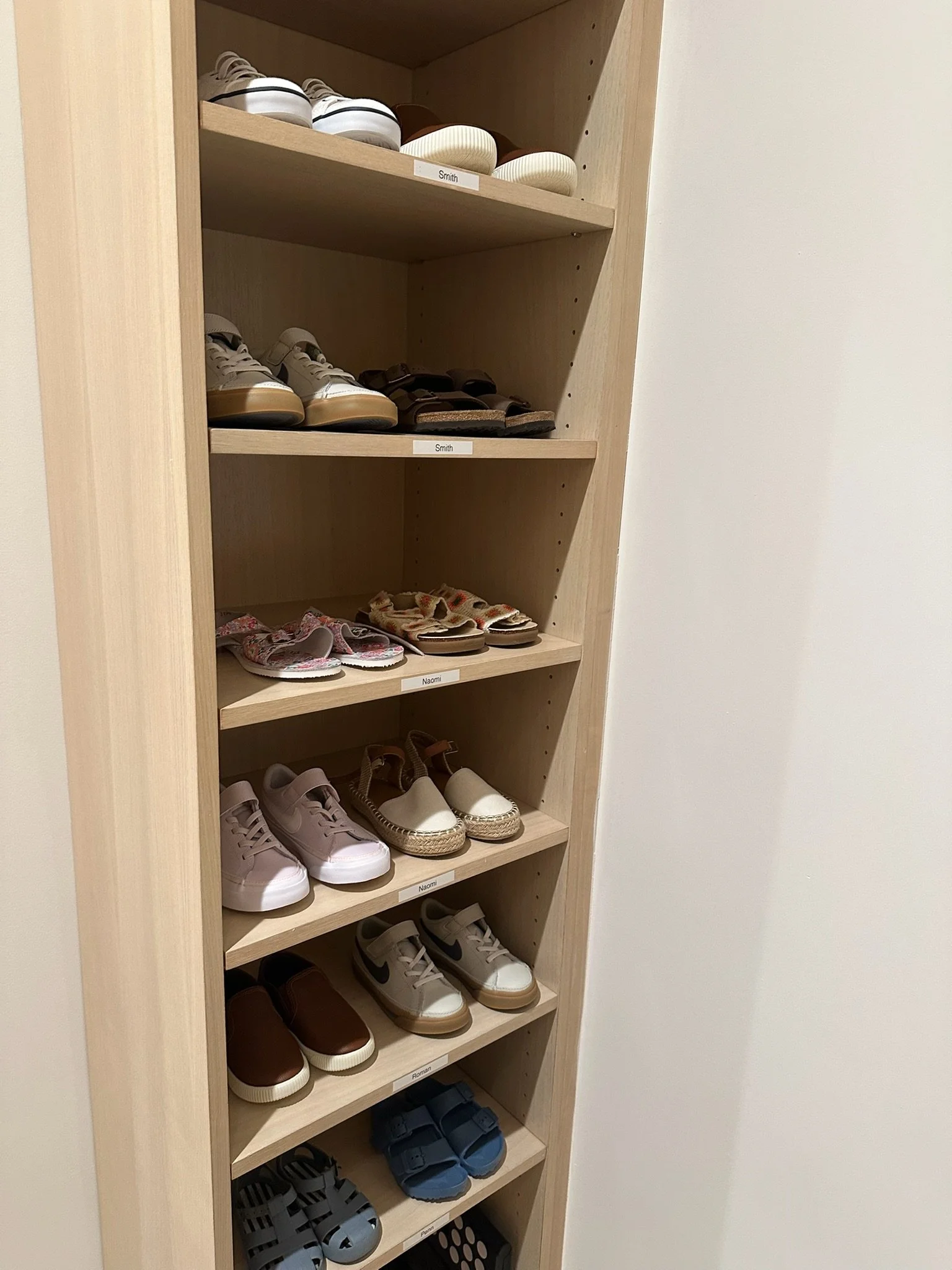 A wooden shoe rack with six shelves holding various pairs of children's shoes, each labeled with names like Smith, Naomi, and Roman.