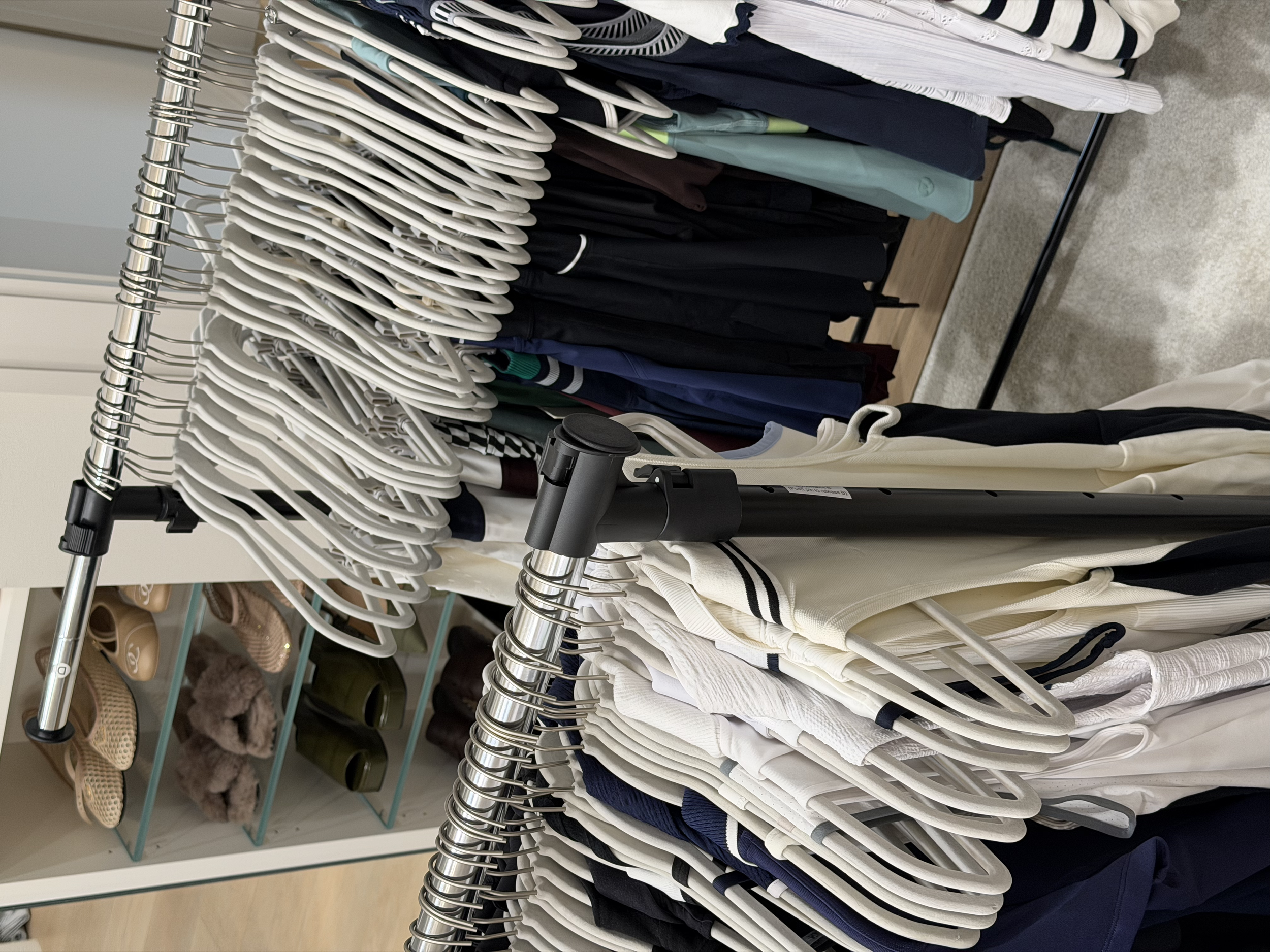 Clothing rack with white, black, and patterned shirts and jackets, and shelves with รองเท้า slippers, fur slippers, and cups in the background.