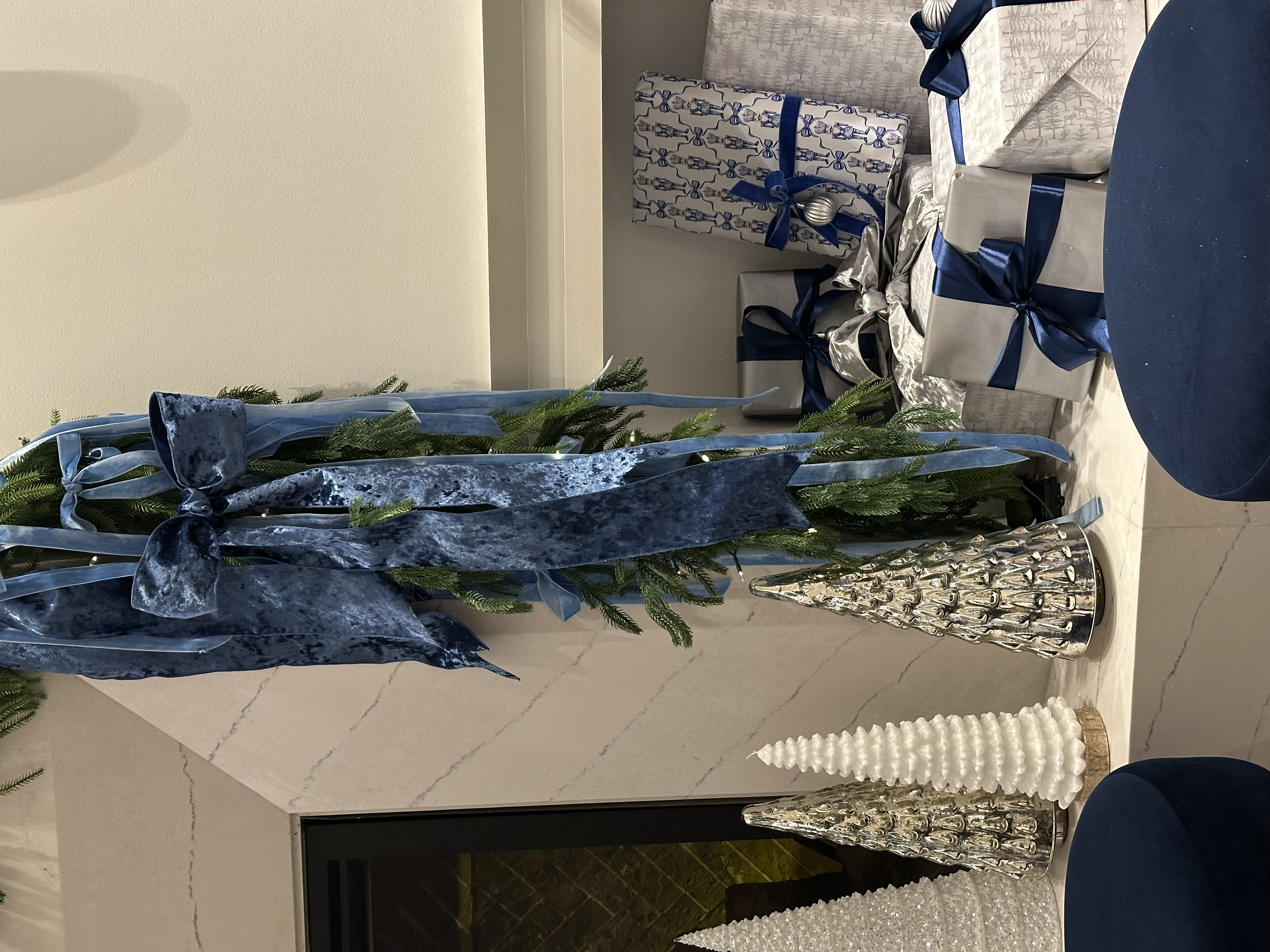 Decorative Christmas display with a small green tree wrapped with blue velvet ribbon, silver and white cone-shaped holiday trees, and wrapped presents with blue ribbons in the background.