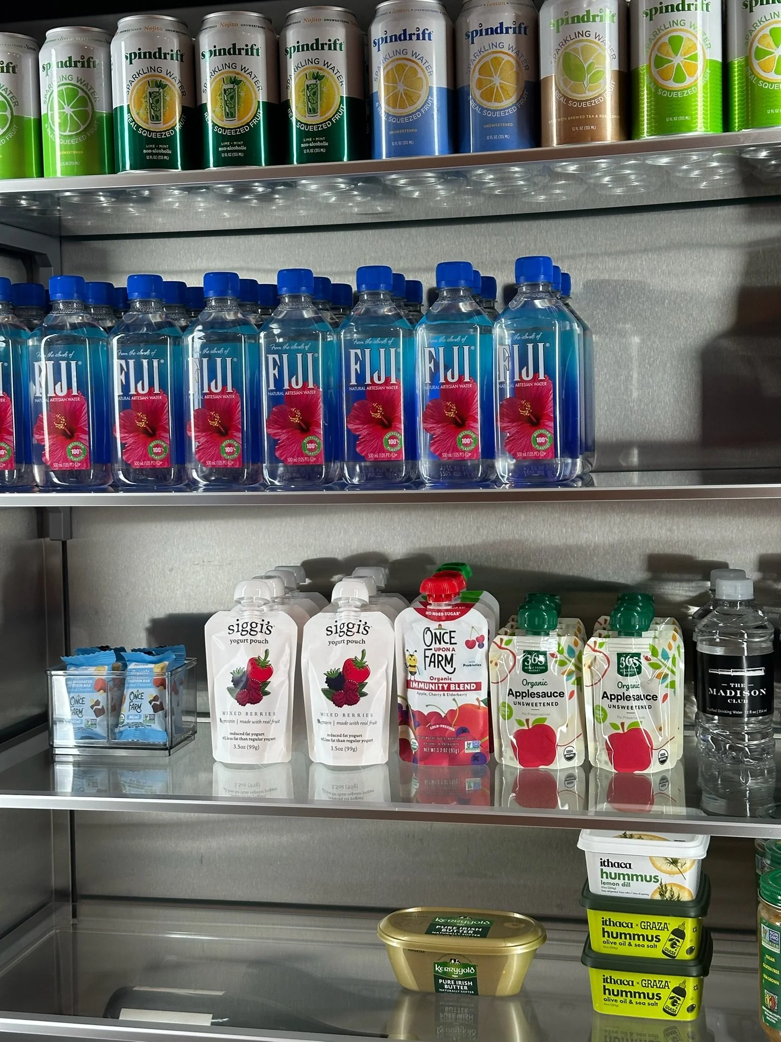 Shelf with various beverages including cans of Spindrift sparkling water, bottles of Fiji water, and cartons of organic apple sauce and yogurt pouches.