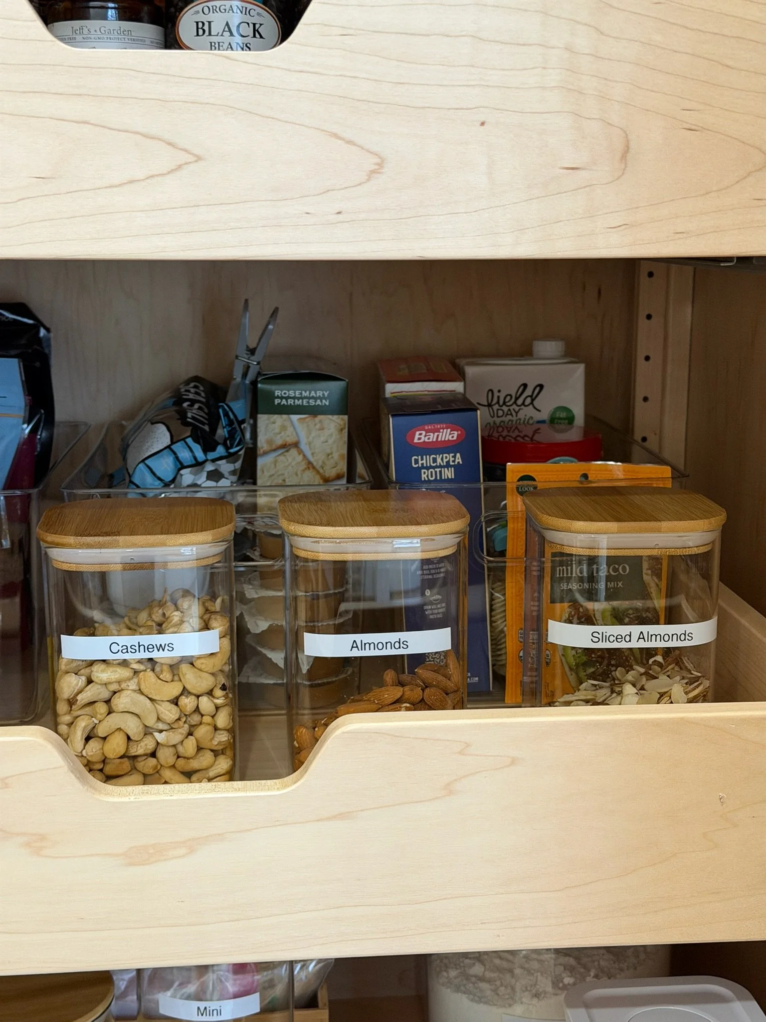 Clear jars with wooden lids containing cashews, almonds, and sliced almonds on a wooden shelf in a pantry or kitchen cabinet.