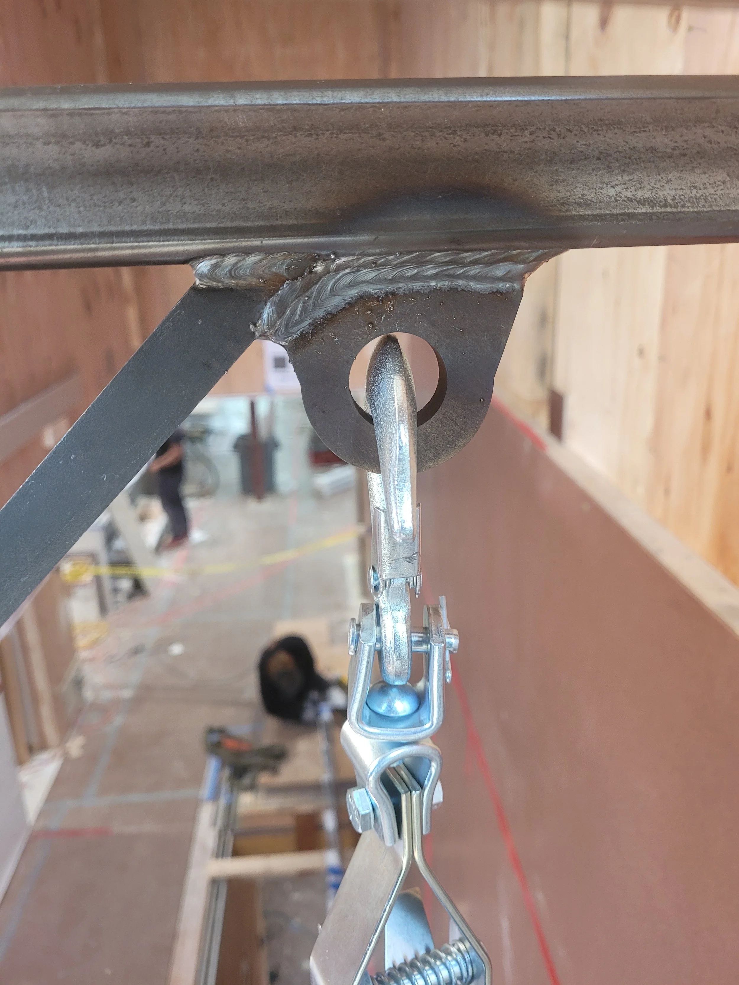 Close-up of a metal hanging wire hook attached to a table in a woodworking shop with a worker and tools visible in the background.
