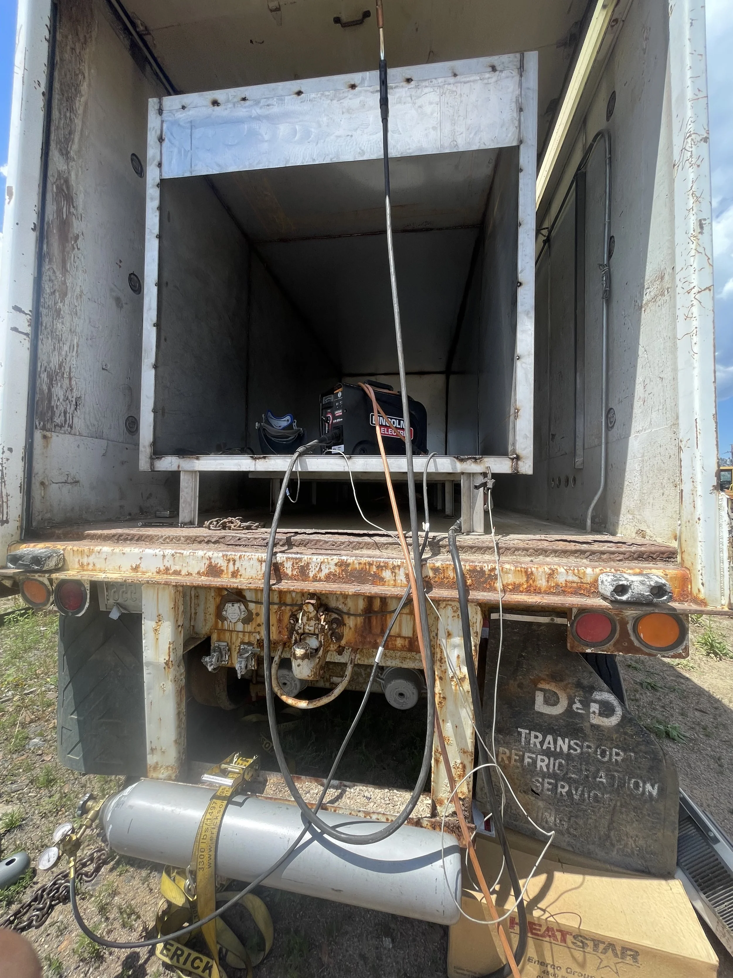 Back of a rusty, white transport truck with an open cargo area containing a portable generator, electrical wires, a small unused propane tank, and a cardboard box labeled 'BEATSTAR' on the ground.