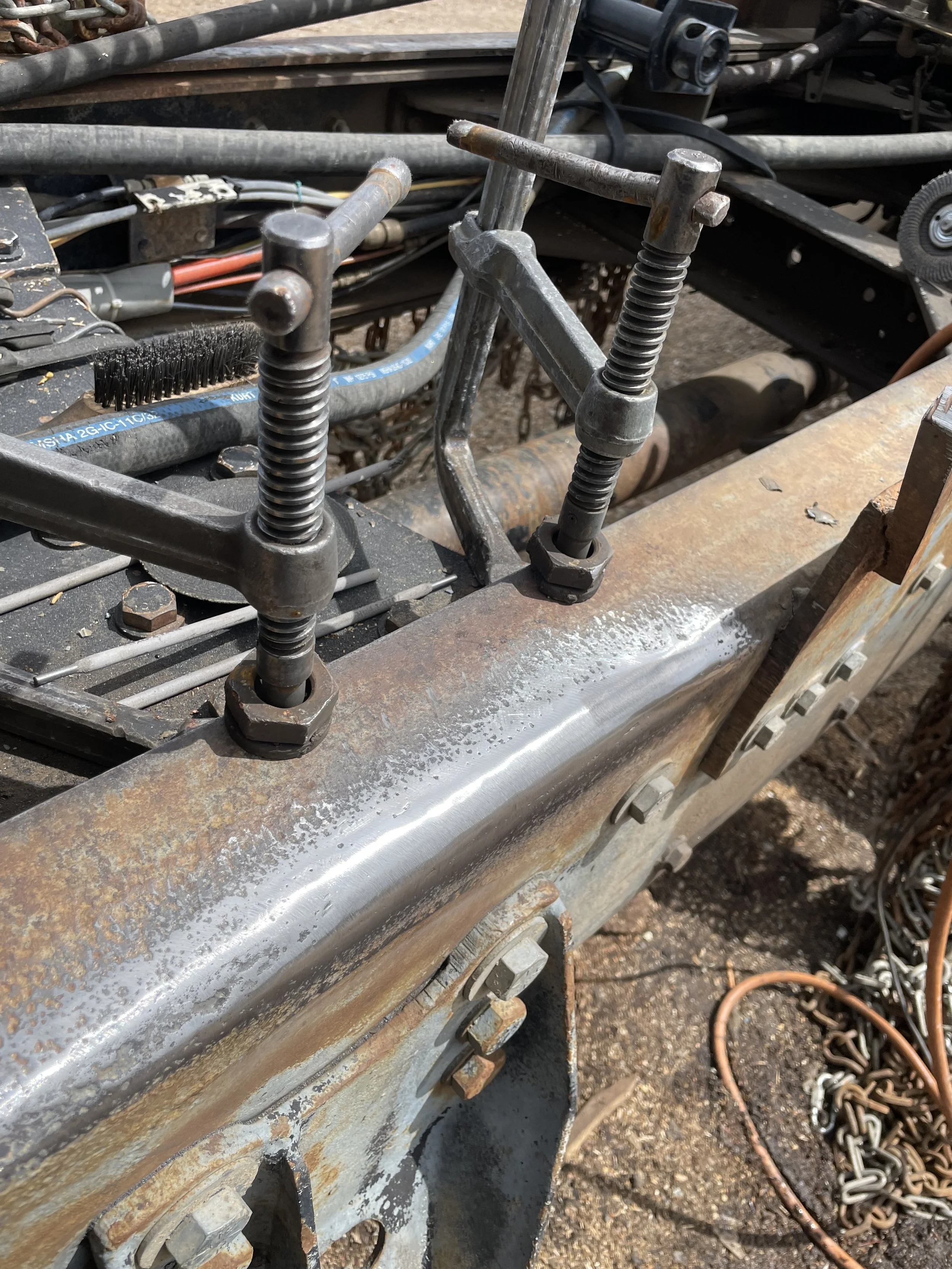 Close-up of a metal piece with two large clamps attached, with bolts and nuts, and rust spots, along with various wires, chains, and mechanical parts in the background.
