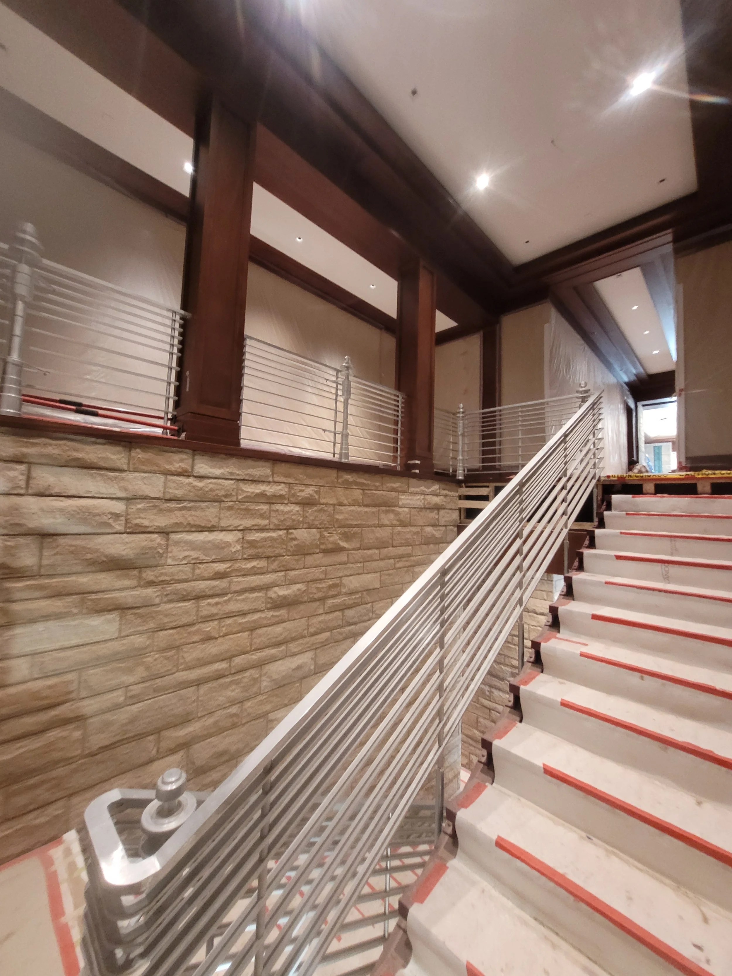 Interior staircase with white steps and a metal handrail, leading upwards with construction tape on the edges, next to a brick wall and wooden beams, with recessed lighting in the ceiling.