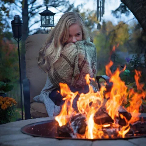 A woman sitting outside near a fire pit, covering her mouth and nose with her sweater, with trees and outdoor lanterns in the background.