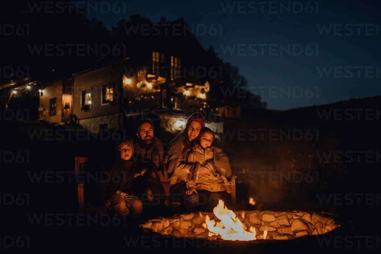 A family of four sitting around a campfire outside at night on a hillside with a house in the background, enjoying the warmth and each other's company.