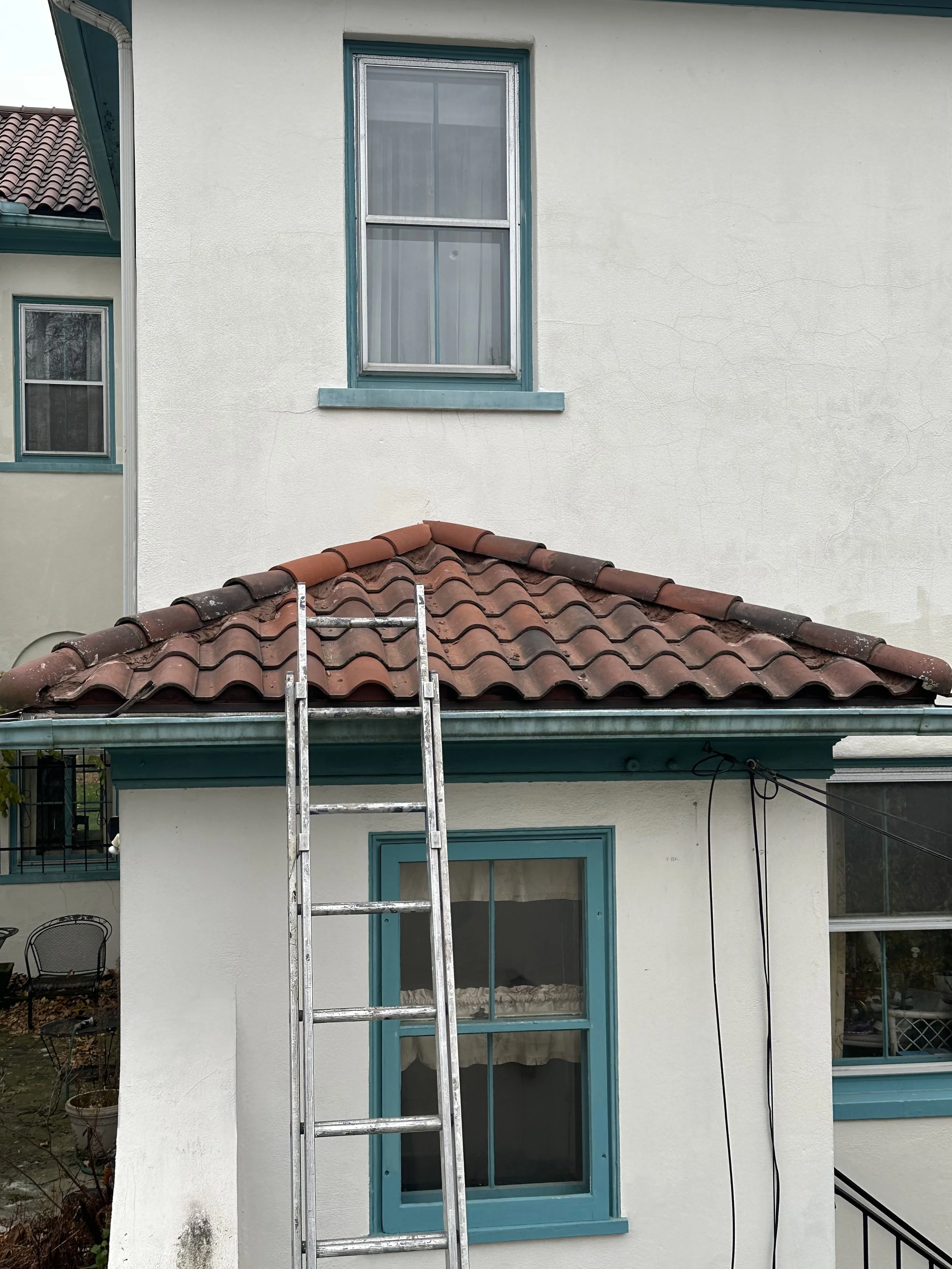 View of a house with a ladder, showing roof tiles, an exterior window, and a white wall with teal trim.