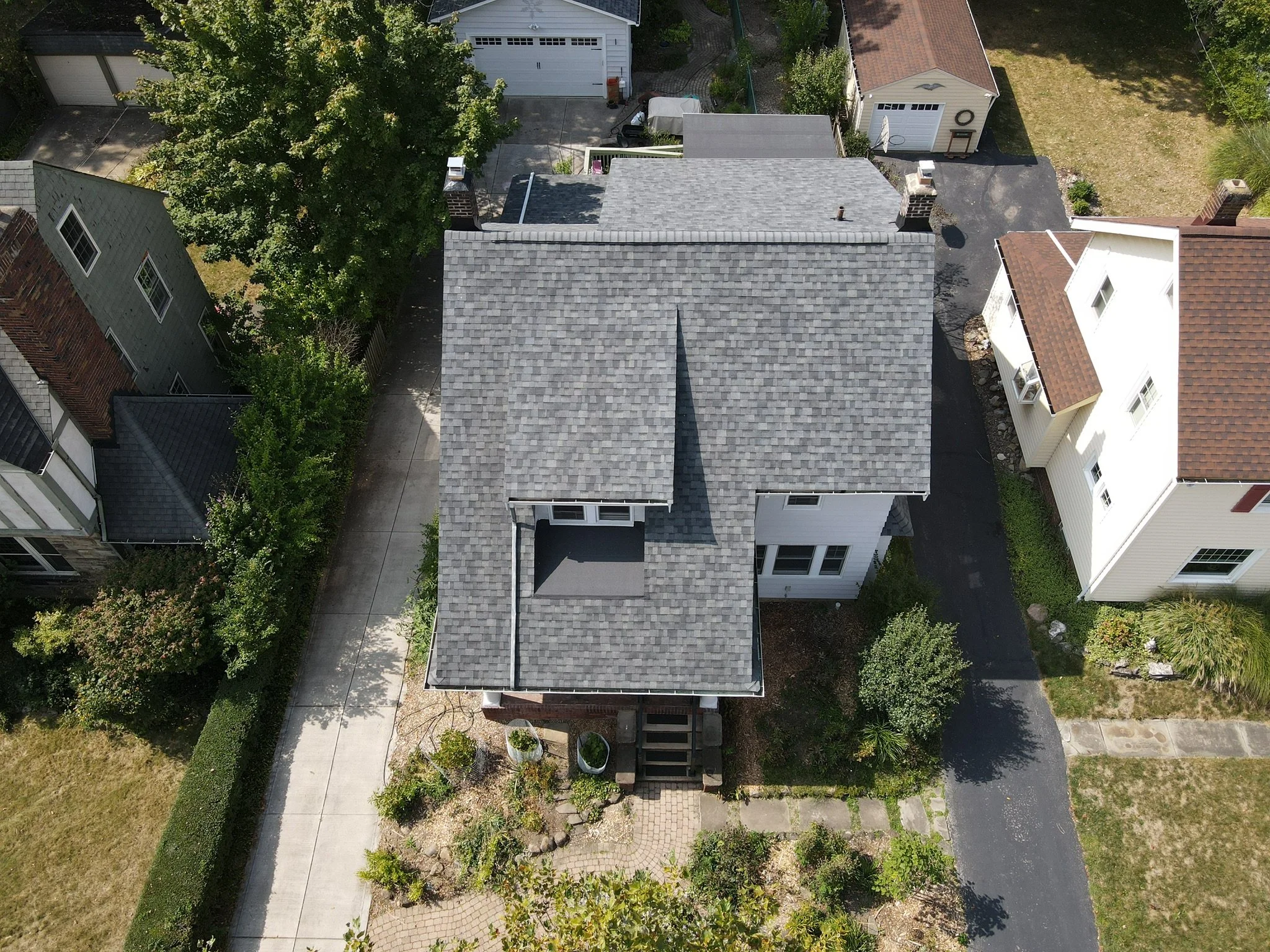 An aerial view of a house with a gray shingle roof, small front porch, and surrounding trees and shrubbery, located in a neighborhood with sidewalks, driveways, and other houses nearby.