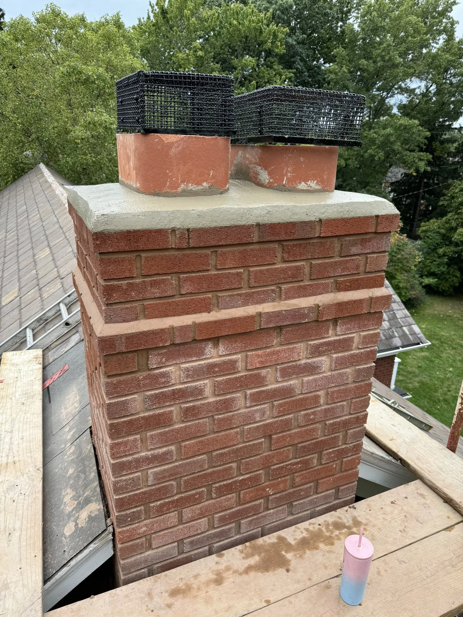 Close-up of a brick chimney on a rooftop with a stack of black plastic grill covers or mats on top, tree-lined neighborhood in the background, and a multicolored candle on the wooden surface in front.