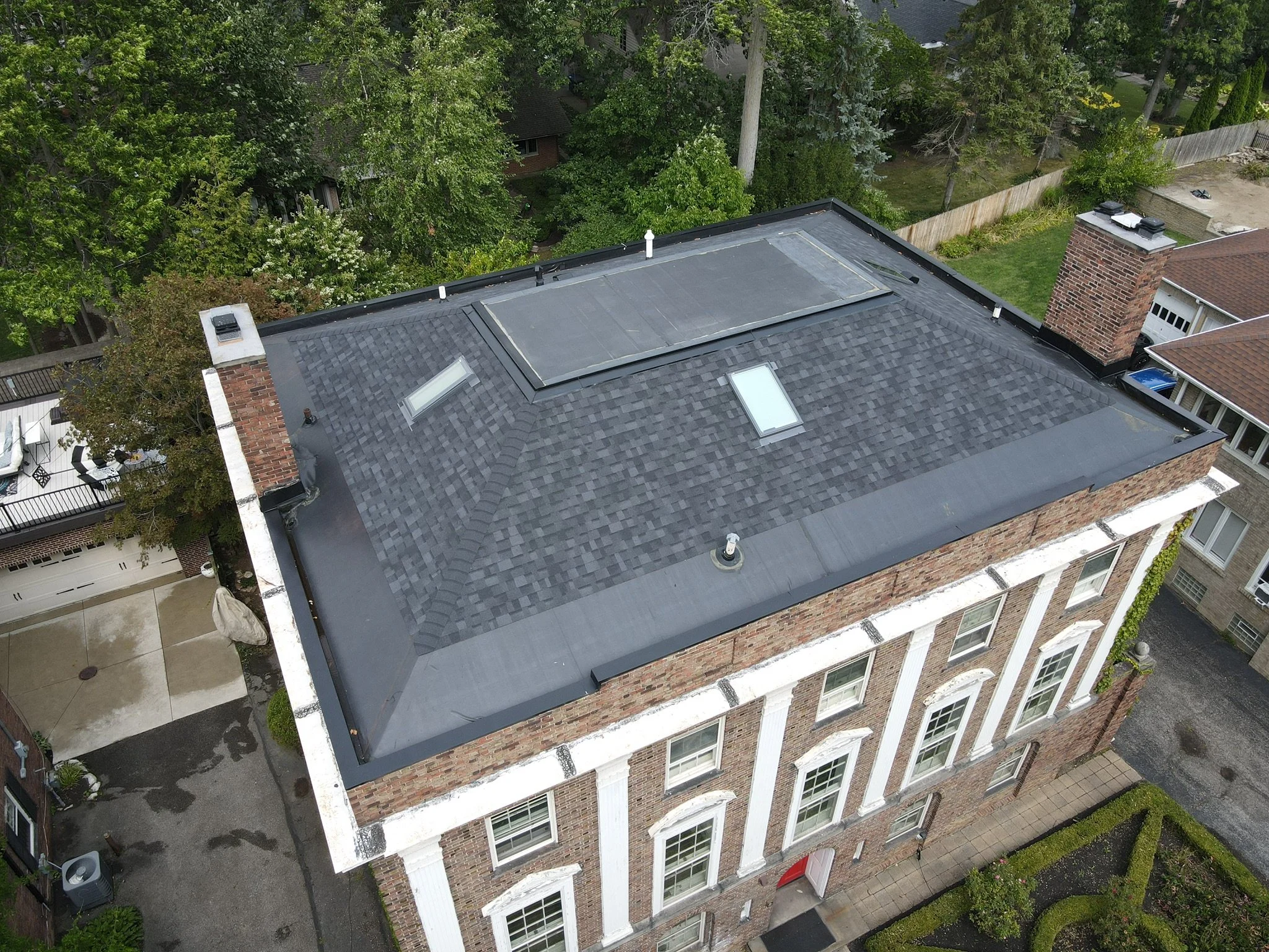 Aerial view of a brick building with a dark-shingled roof. The roof features two skylights and a larger, raised solar panel. Surrounding the building are trees, neighboring houses, a small yard, and a parking area.