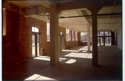 Empty industrial space with large windows, brick walls, and wooden beams, illuminated by sunlight.