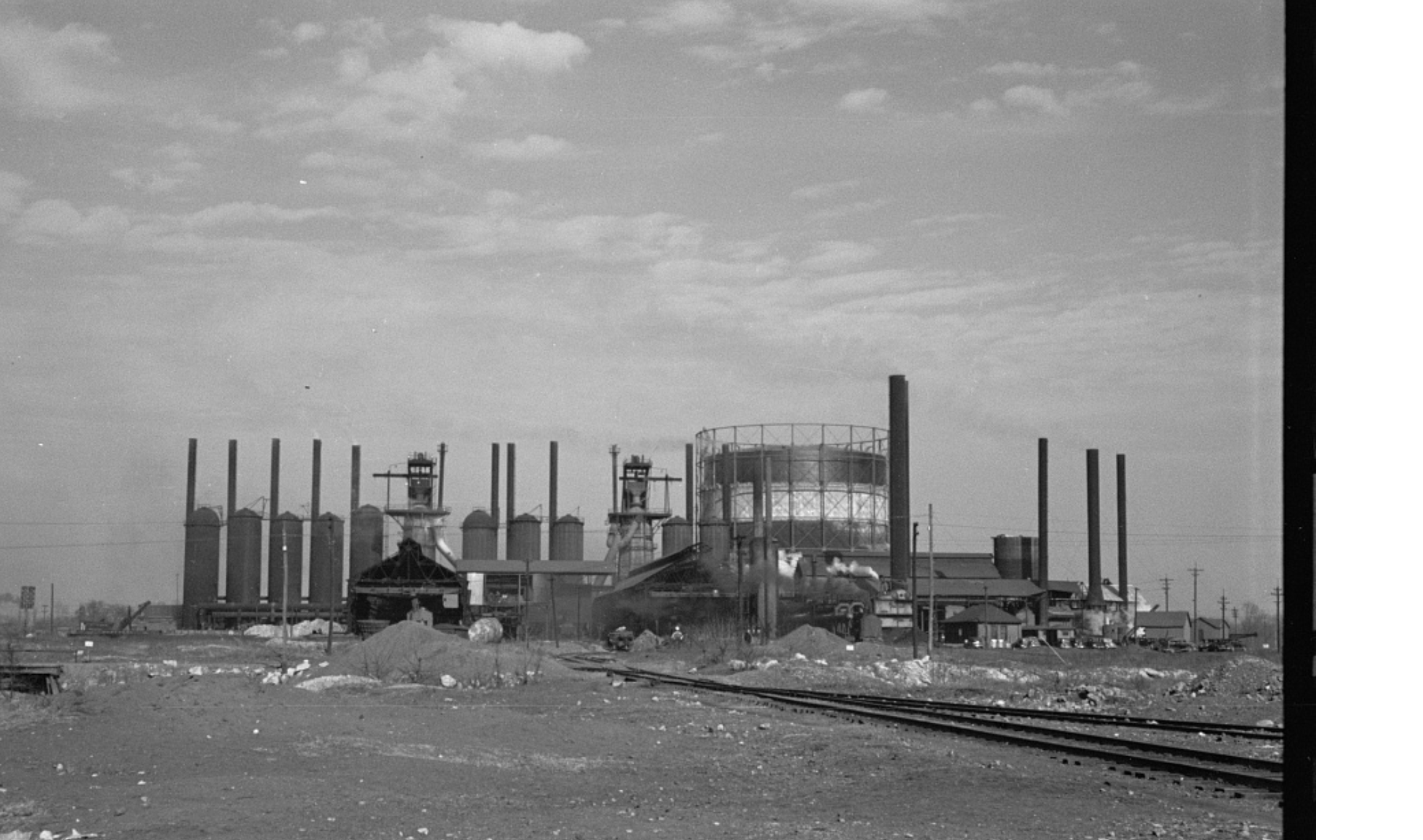 A black and white photo of an industrial power plant with multiple smokestacks, cooling towers, and utility structures. The foreground shows a dirt area with scattered debris and a set of railroad tracks.
