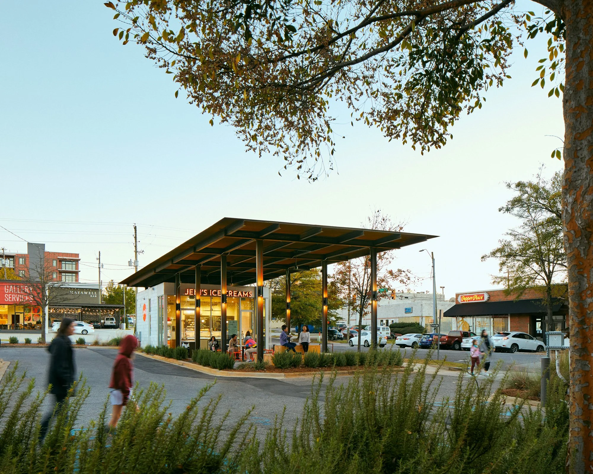 A small outdoor eating area with a modern roof structure, surrounded by parked cars and trees, in a commercial district during early evening.