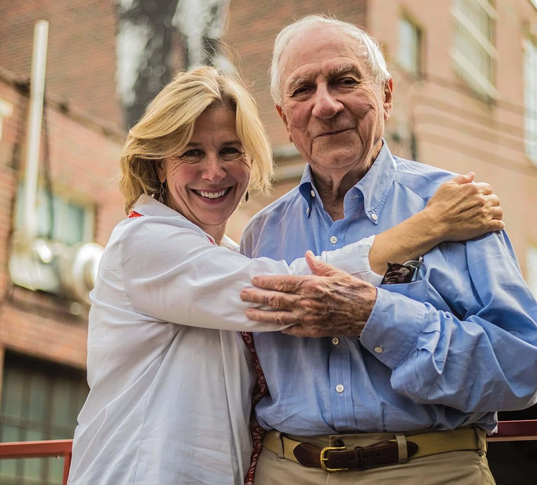 An older woman and man are embracing and smiling together outdoors in front of brick buildings.